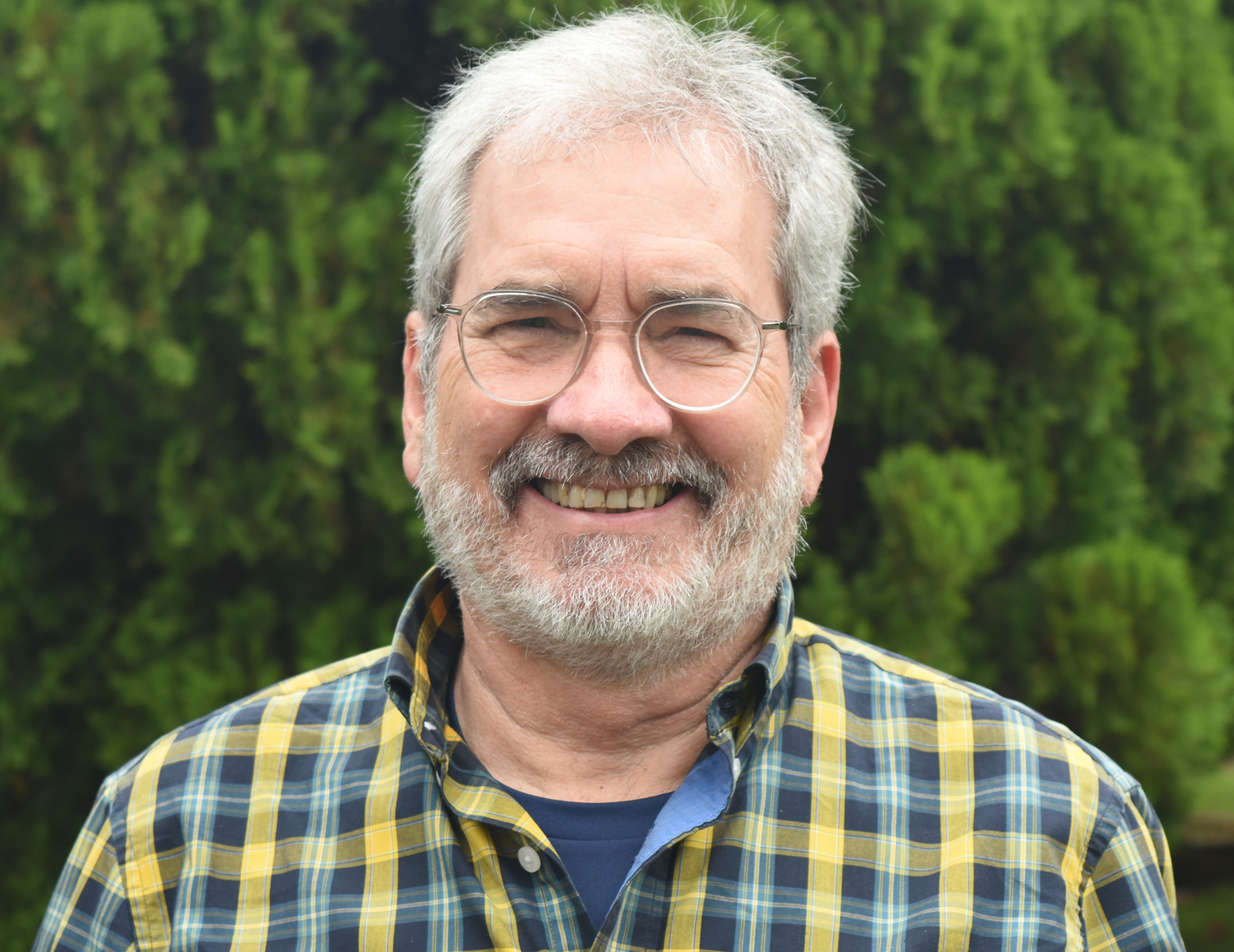 Smiling older man with gray hair, glasses, and a beard, wearing a yellow plaid shirt, outdoors with green trees in the background.