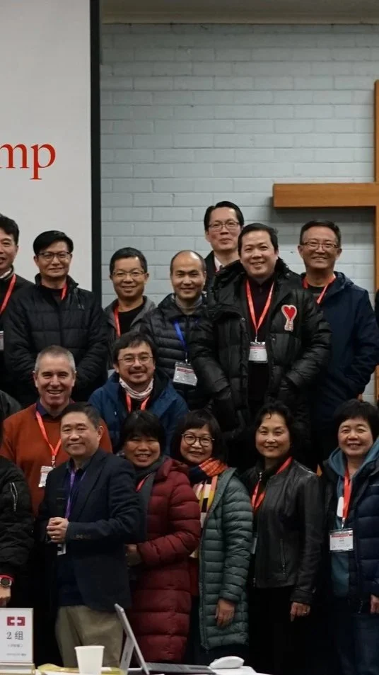 Group of people, mostly men, smiling and wearing winter jackets with red lanyards, standing indoors in front of a light gray brick wall.