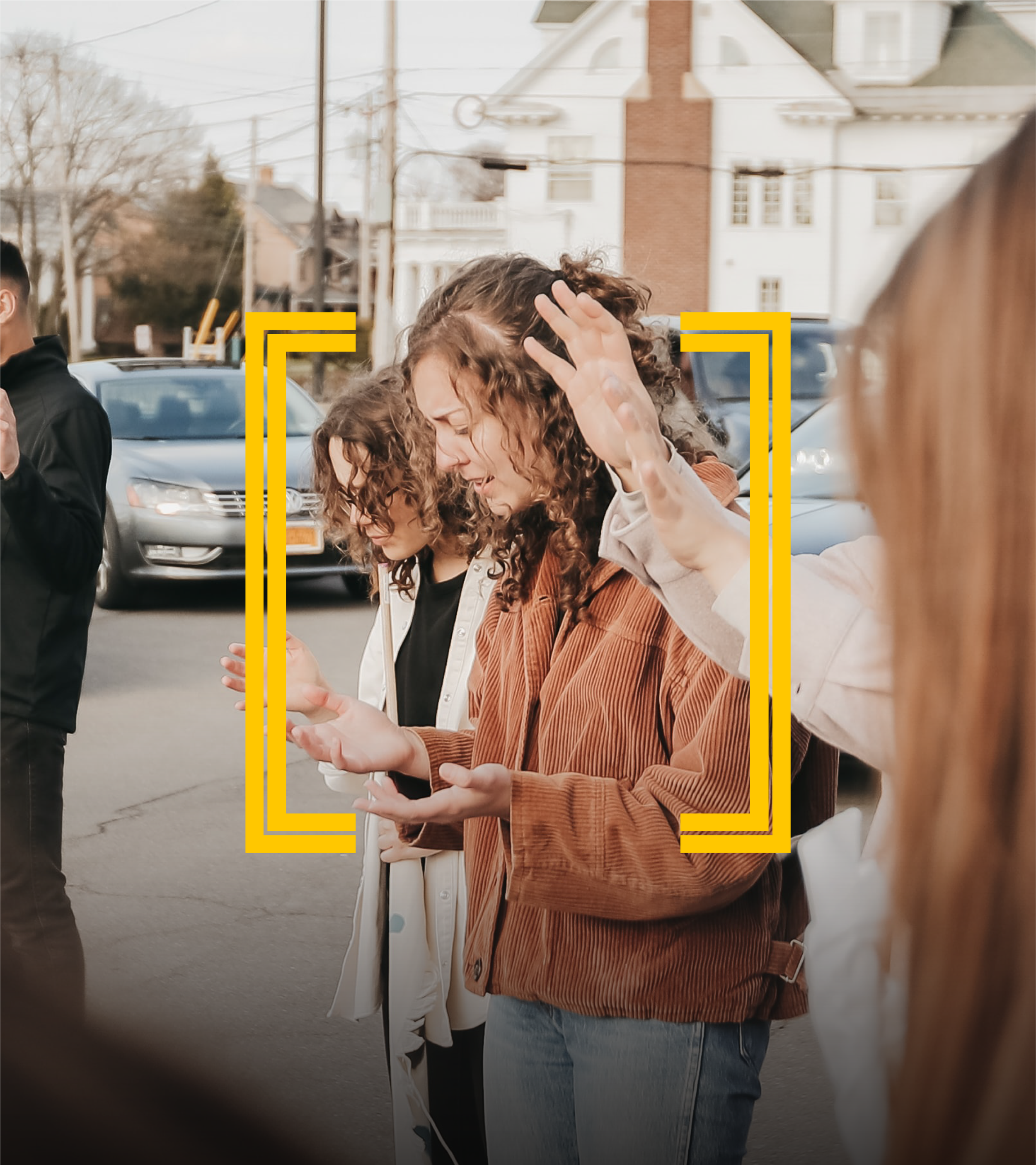People talking and interacting outdoors on a city street during daytime with cars and houses in the background.