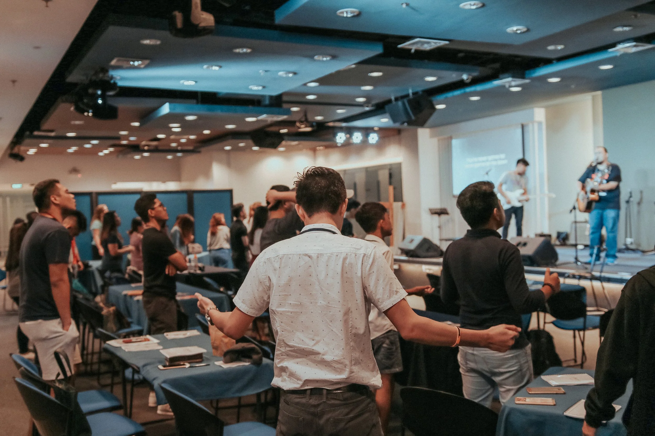 A large group of people standing in a conference room, listening to two musicians playing guitars on a stage at the front of the room.