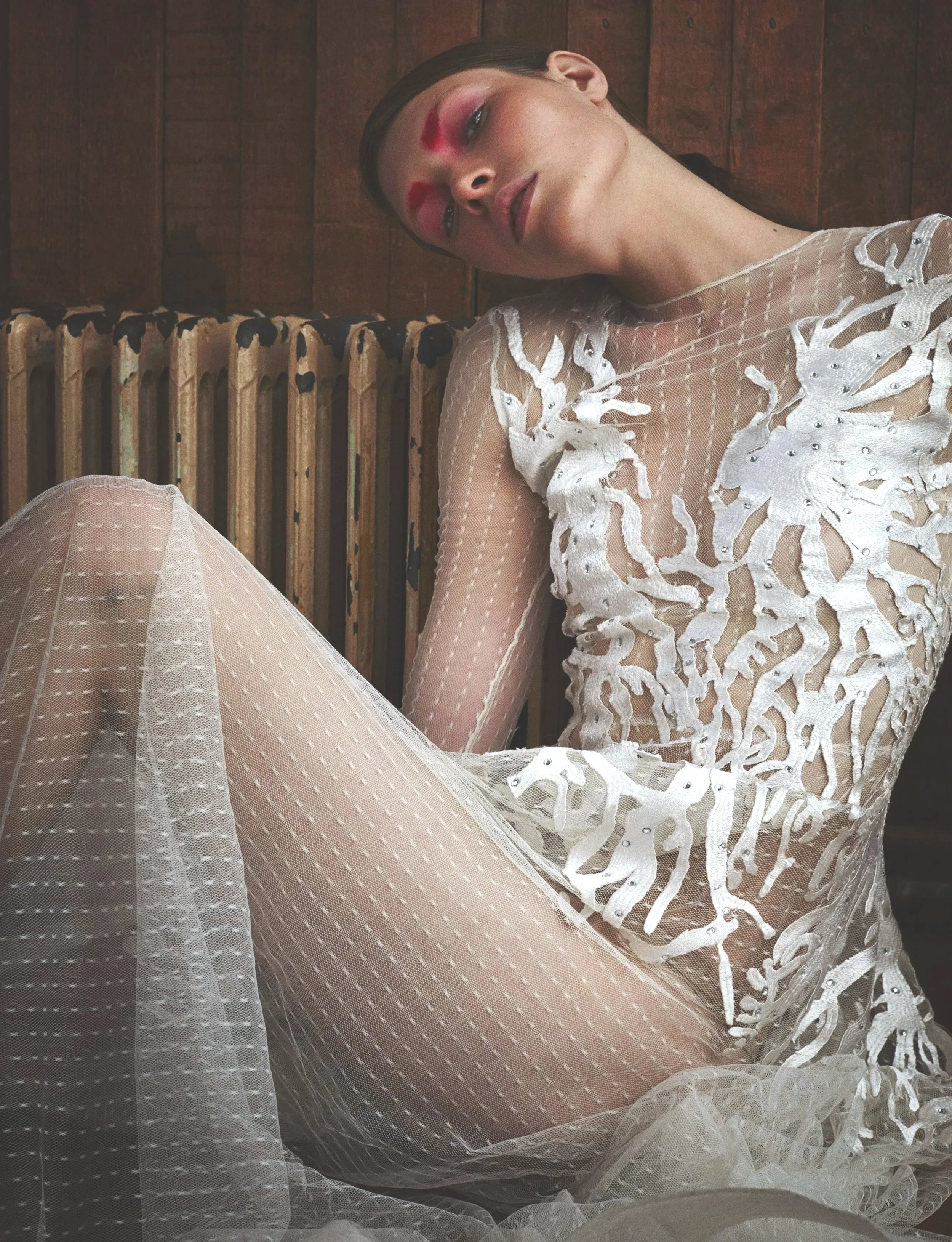 Woman with bright pink eyeshadow and a sheer mesh white dress sitting down.
