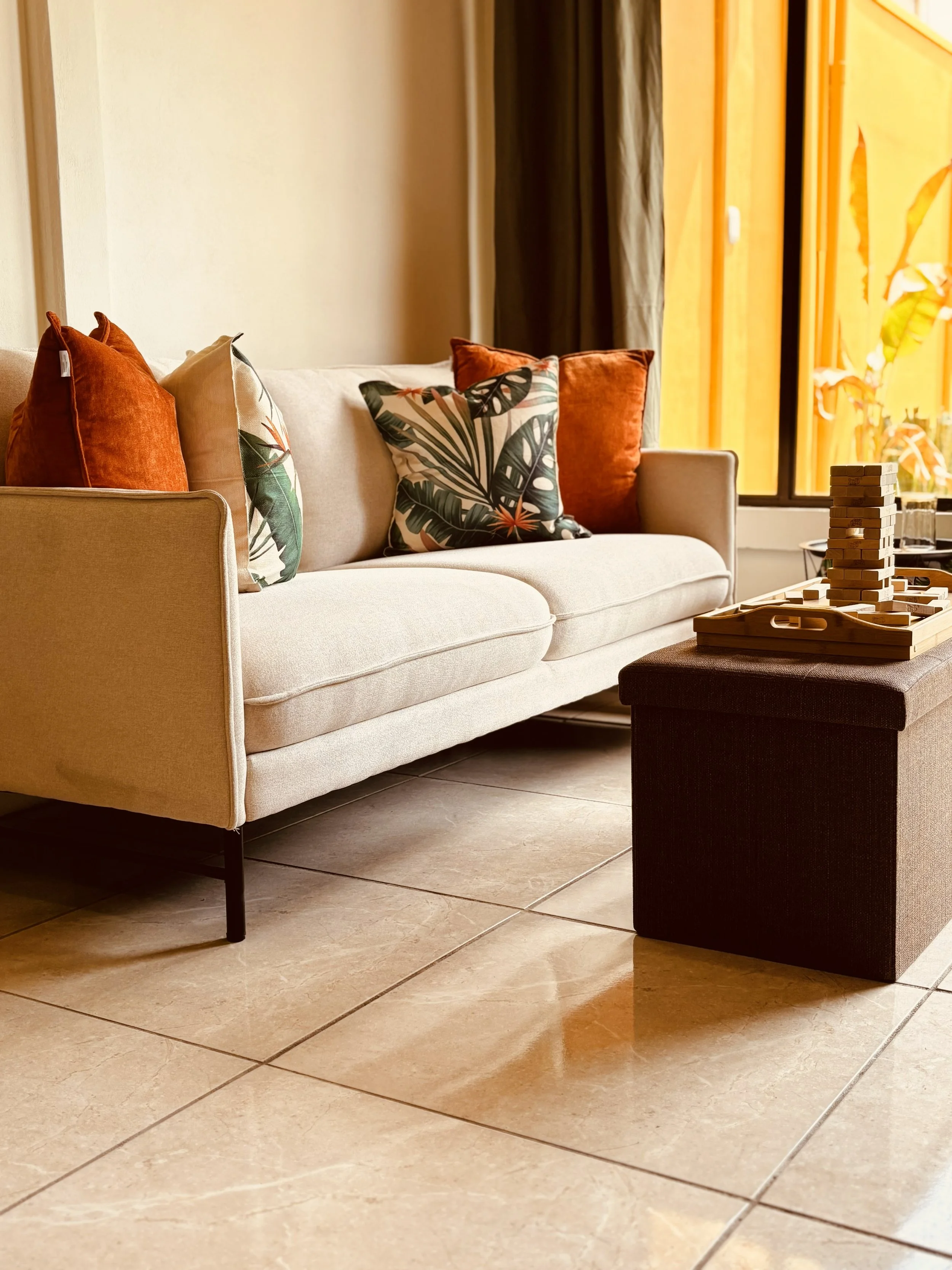 A beige couch with orange and tropical-patterned throw pillows, located in a room with tiled floor and a window with yellow curtains.