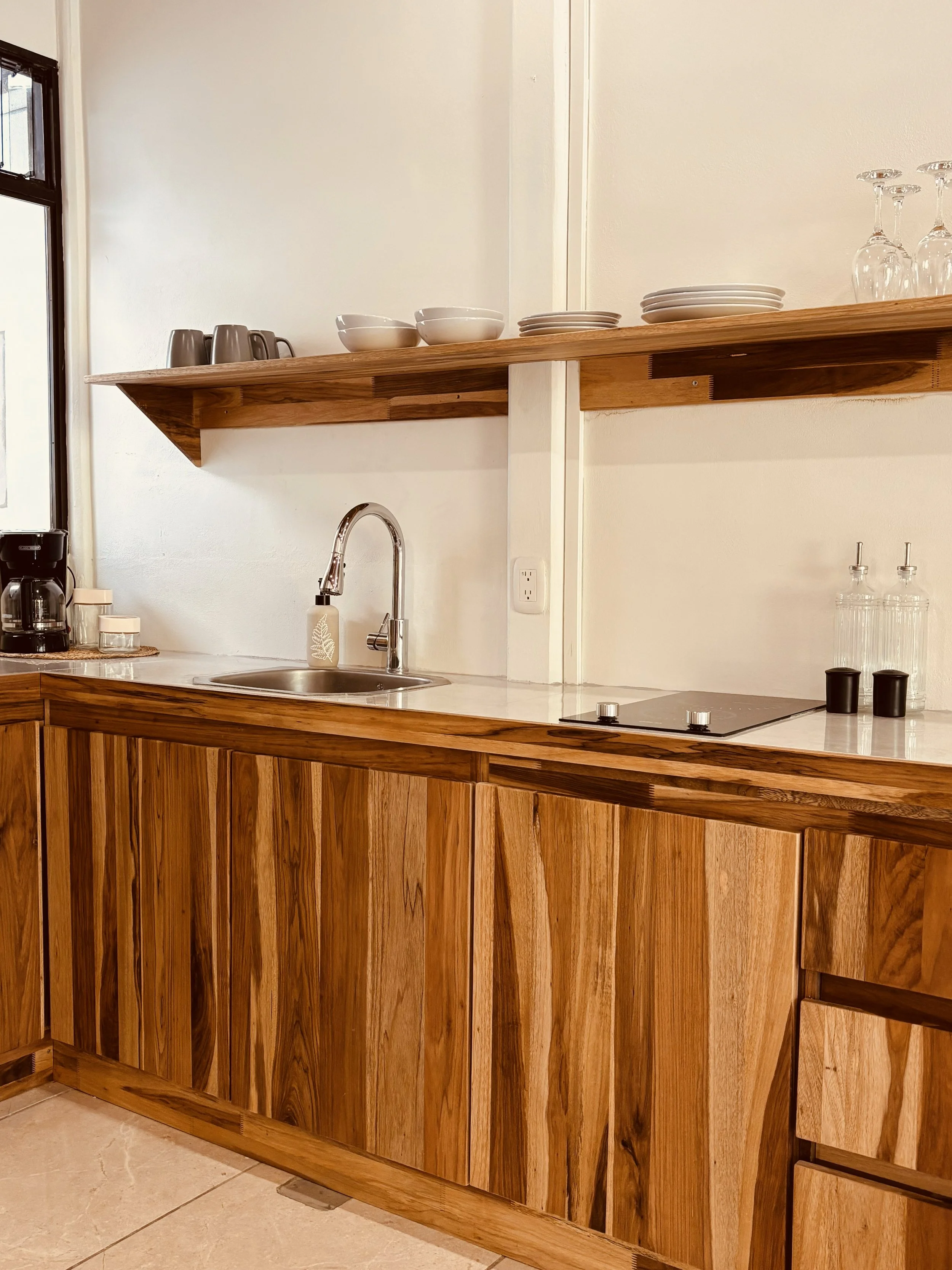 Wooden kitchen counter with a sink, a coffee maker, glasses, and plates on a shelf.