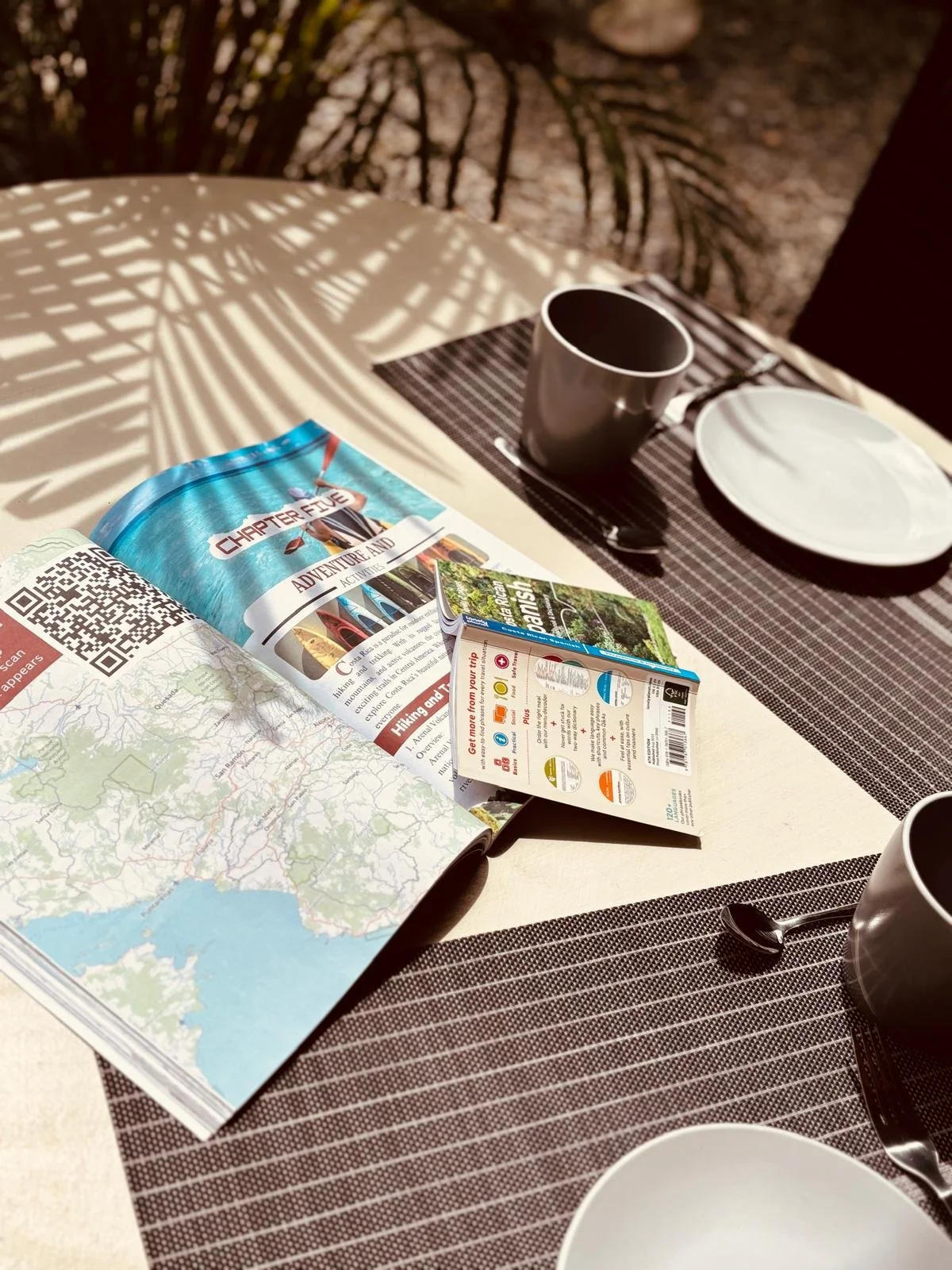 Table with a travel magazine, a map, a coffee mug, a white plate, a spoon, and a brochure, with shadows of plant leaves cast onto the table.