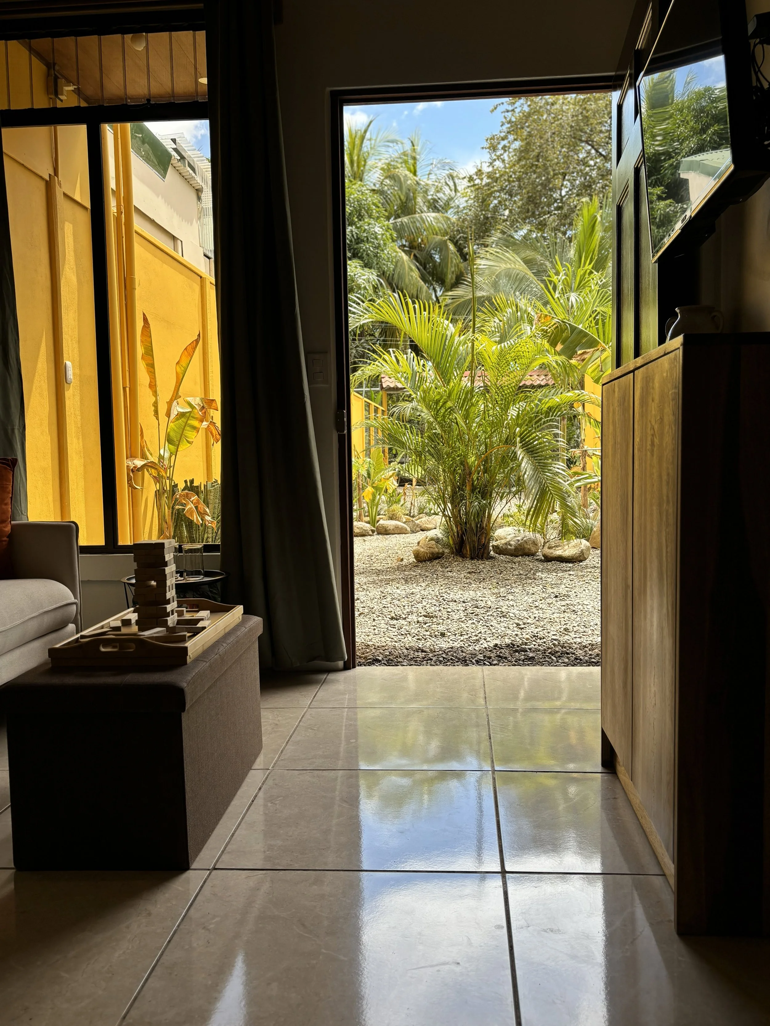 Interior view of a room with a sliding glass door open to a garden with lush green plants and palm trees. Inside, there is a beige upholstered sofa, a wooden cabinet, and a small table with a game or puzzle on it. The floor is tiled and reflects sunl