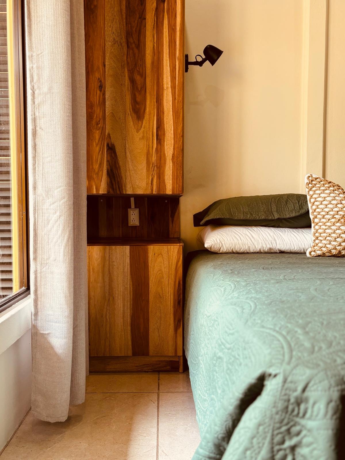 A bedroom corner with a bed, pillows, a wooden wall-mounted cabinet, a black adjustable lamp, a power outlet, beige curtains, and a window with wooden blinds.