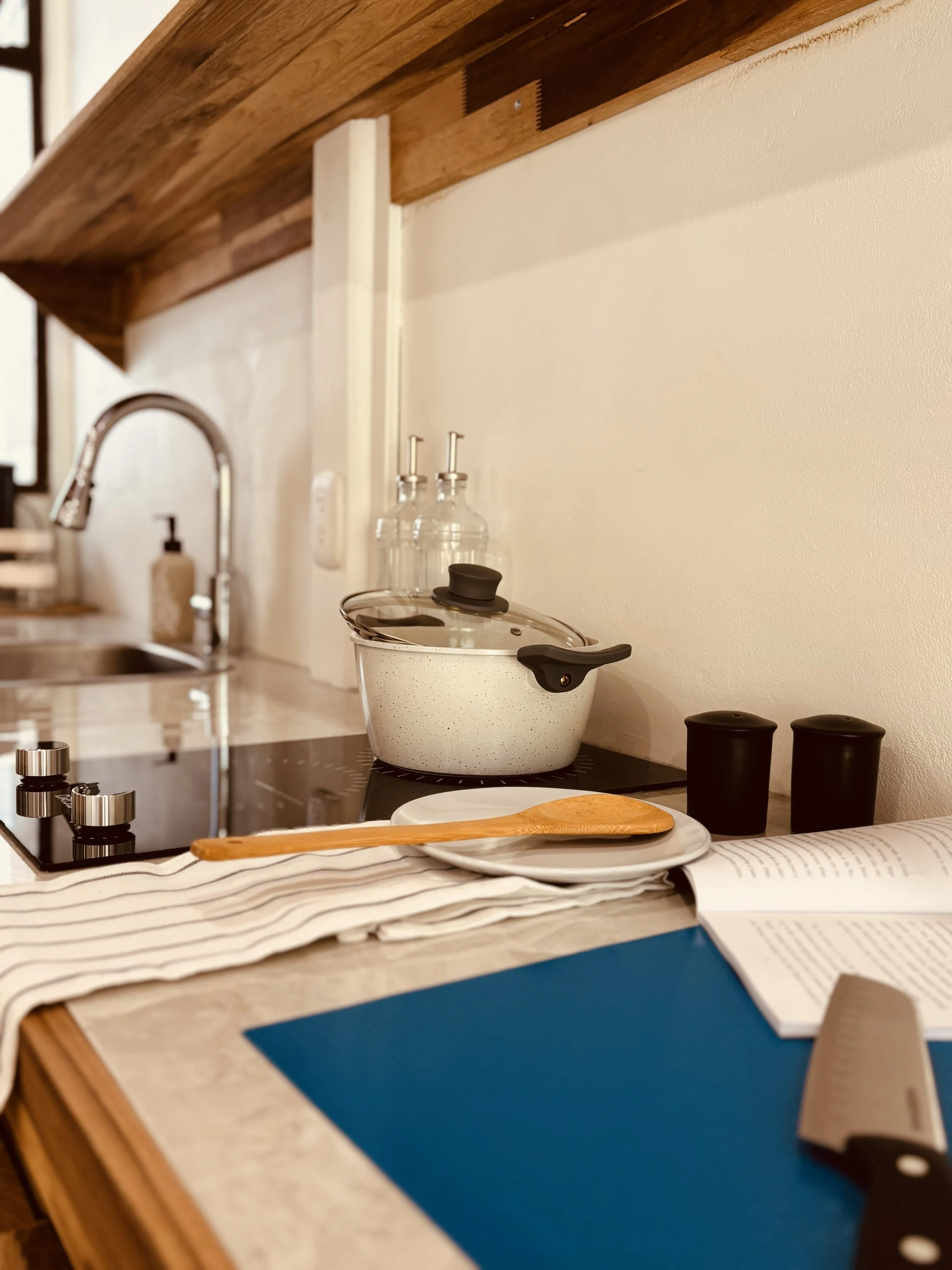 Kitchen counter with black stovetop, white speckled pot with black lid, wooden spoon on plate, open book, and blue cutting mat