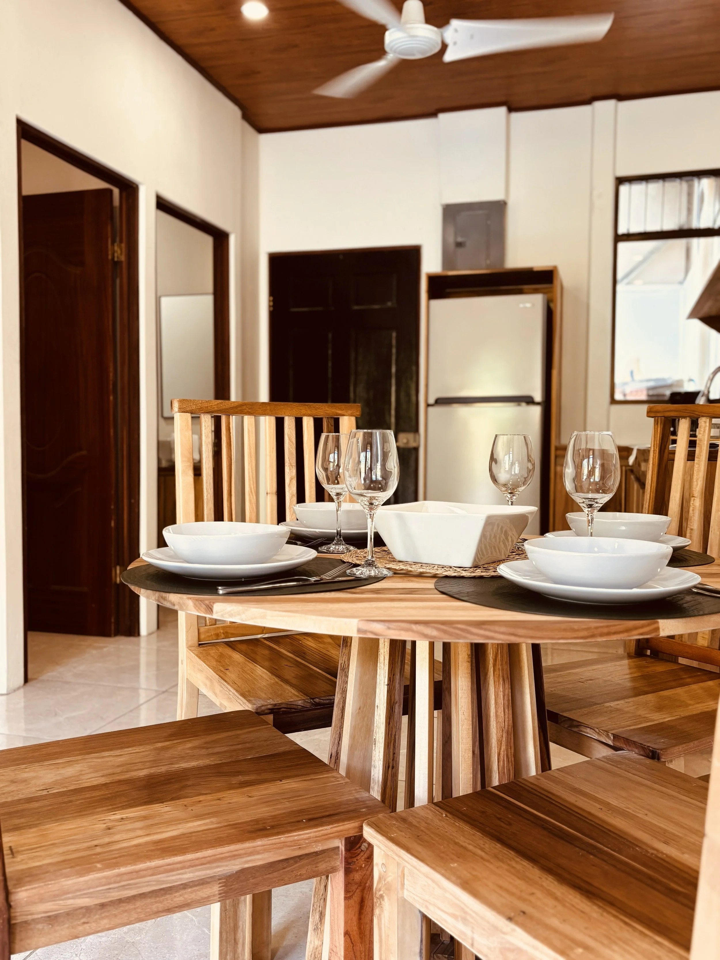 Kitchen and dining area with a wooden table set with white plates, bowls, and wine glasses, with wooden chairs around it. In the background, stained wood doors, a refrigerator, and a window are visible under a wooden ceiling with a ceiling fan.