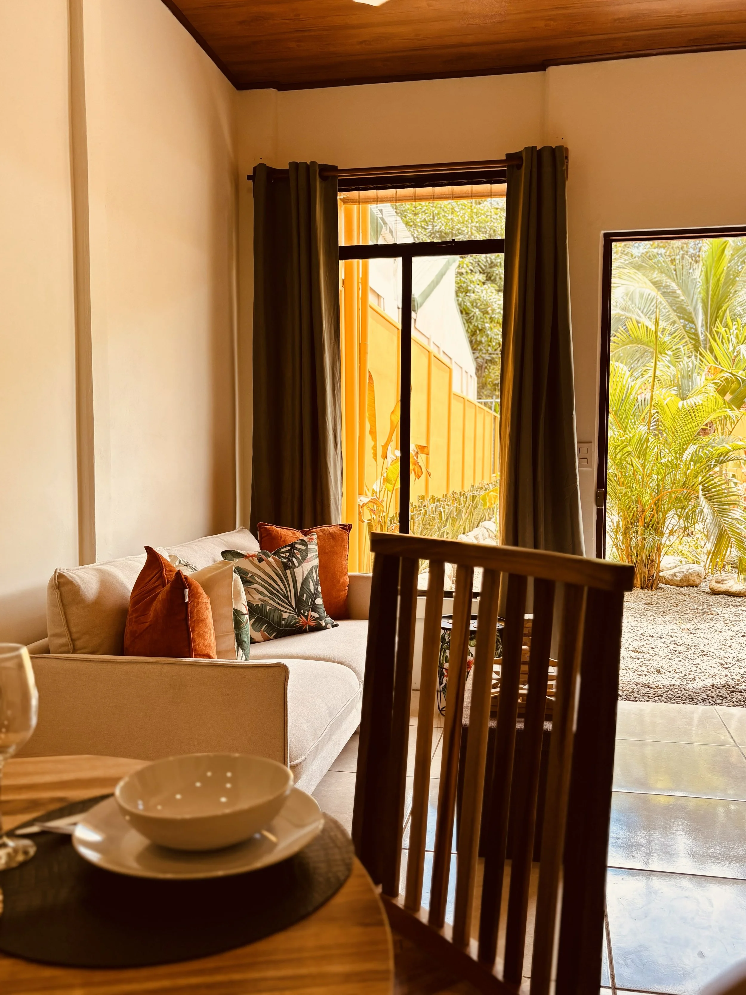 A cozy living room with a beige sofa decorated with colorful pillows, between open patio doors showing a garden with lush greenery and a tall yellow fence, during daytime.