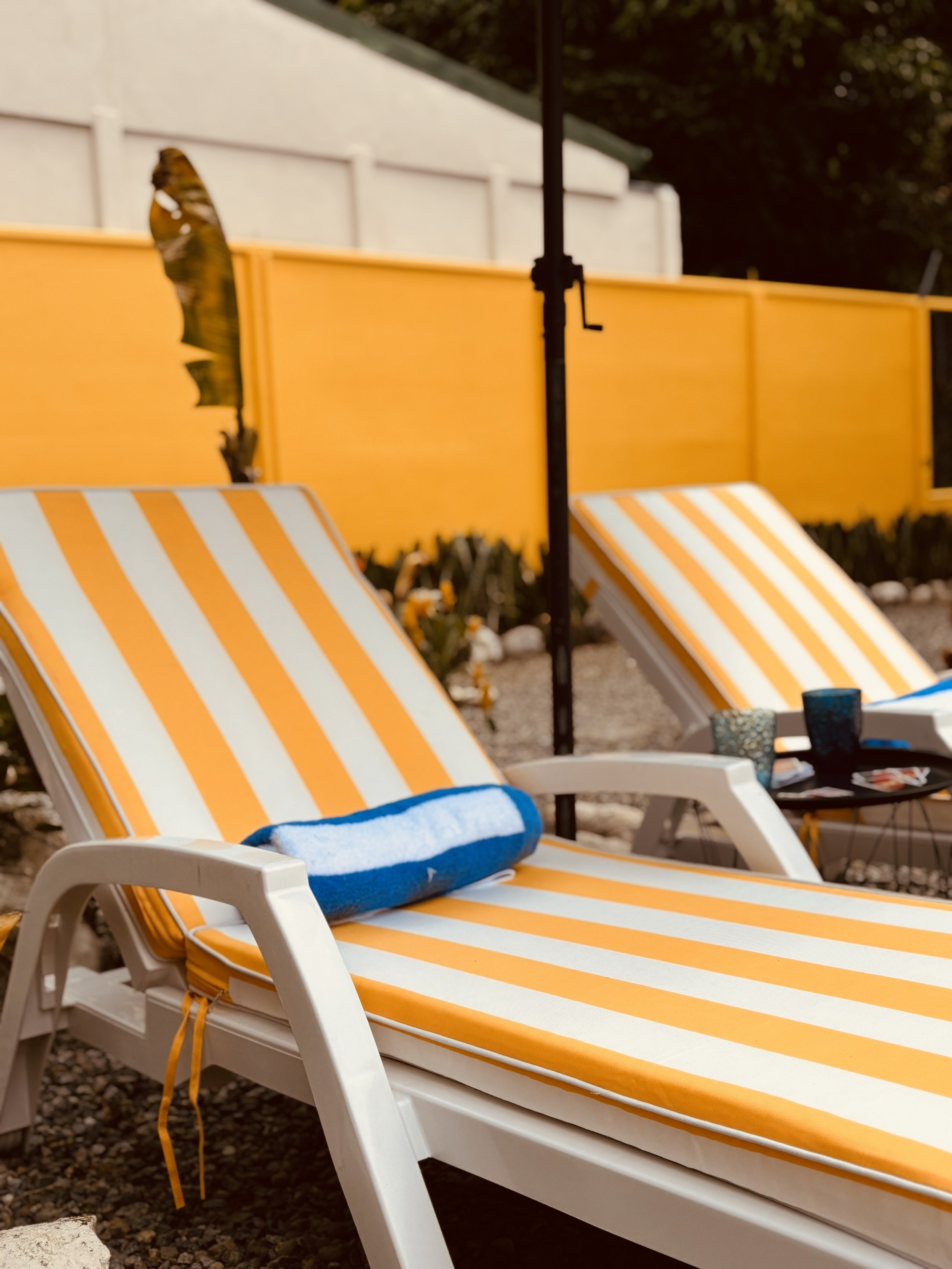 A poolside scene featuring a yellow and white striped lounge chair with a folded blue and white towel on it. In the background, there is a yellow fence, a white fence, a black patio umbrella pole, and some plants and rocks at ground level.
