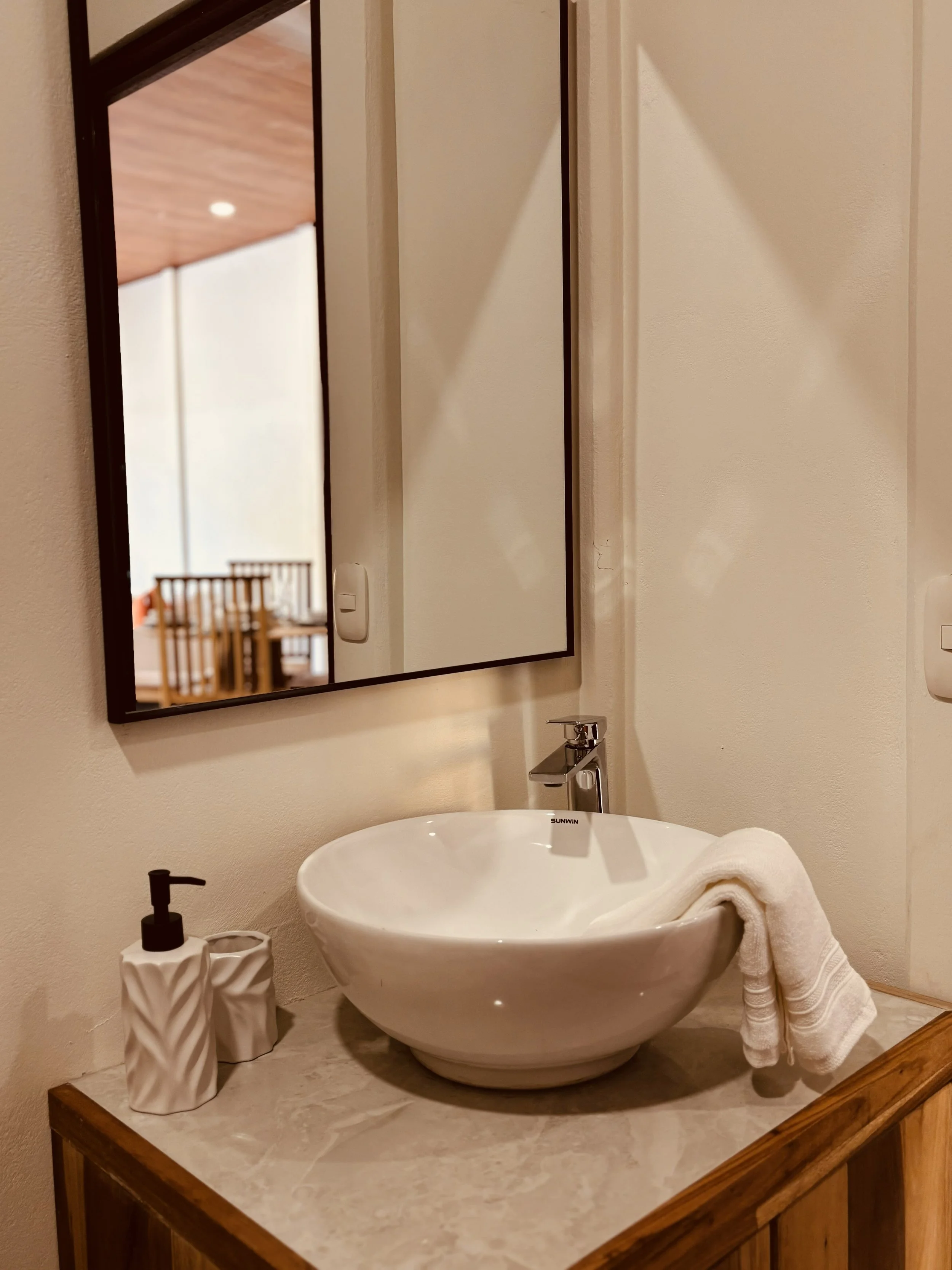 A bathroom sink with a towel on the counter, a soap dispenser, a mirror, and a window showing a wooden ceiling and part of a dining area with chairs.