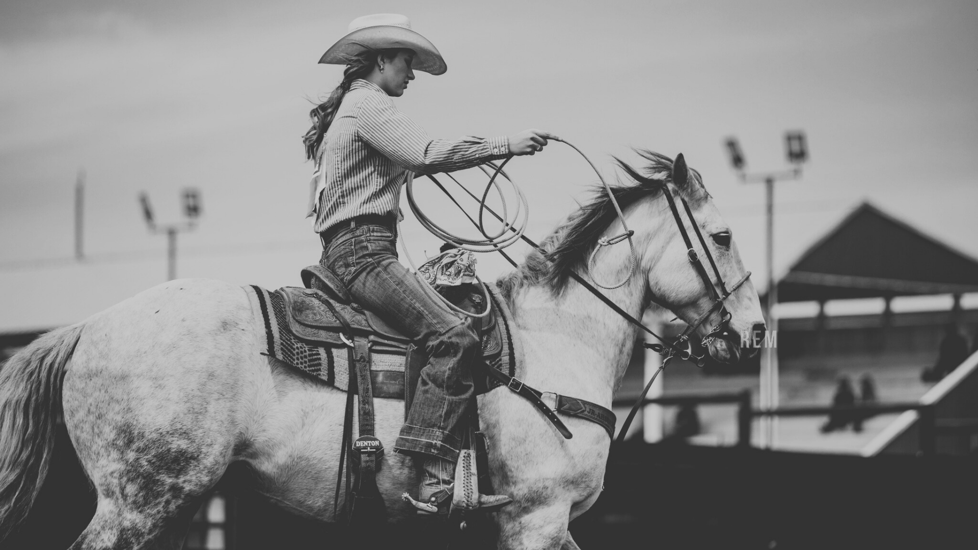 A woman riding a horse, wearing a cowboy hat and striped shirt, holding a coiled rope.