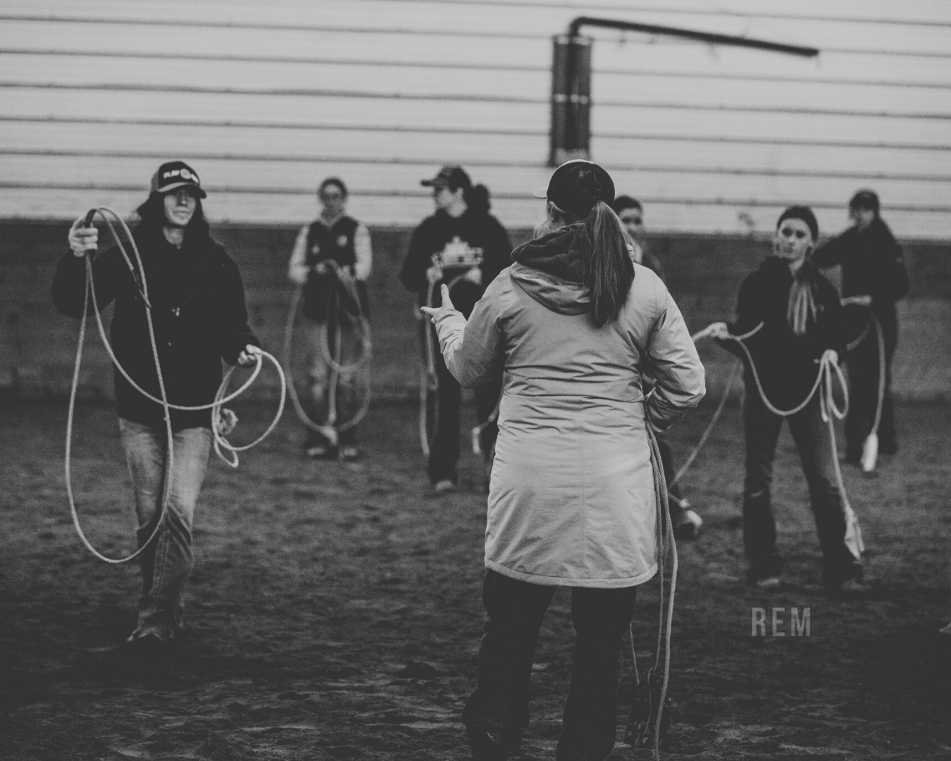 A woman in a jacket and hat leading a group of women practicing Breakaway Roping skills indoors. The women are holding ropes and appear to be in a training session, with some looking at the instructor and others focusing on their own practice.