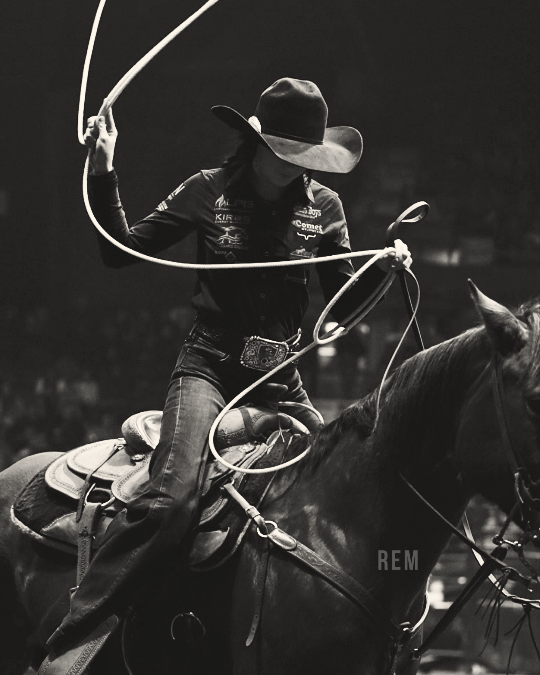 Black and white photo of a female Breakaway Roper on horseback, wearing a wide-brimmed hat, western shirt, jeans, and belt with a large buckle, swinging her rope in the air.