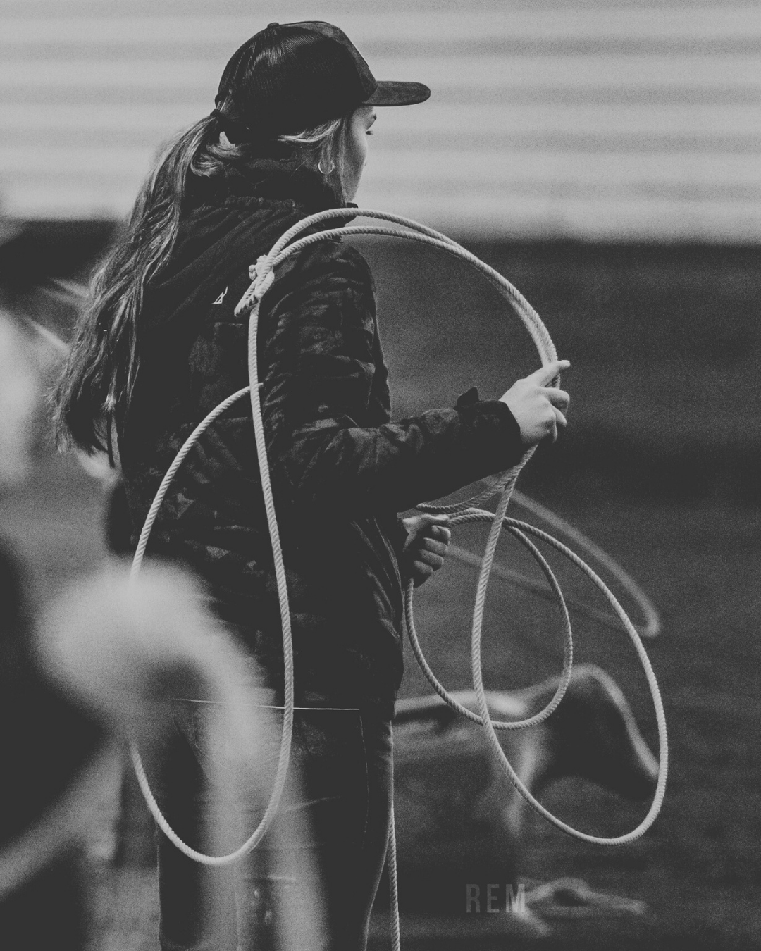 A woman wearing a cap and a dark jacket, holding a rope, preparing to rope the plastic calf from the ground.