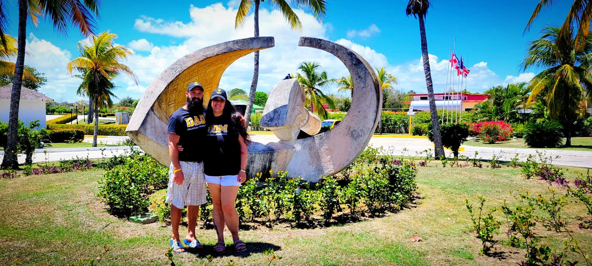 A man and woman standing together in front of a large public sculpture at a park with palm trees, colorful flowers, and flags, on a sunny day.