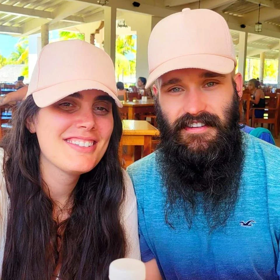 A smiling woman and a man with a beard, both wearing light pink caps, seated at a restaurant with wooden tables and chairs, tropical scenery visible through open windows in the background.