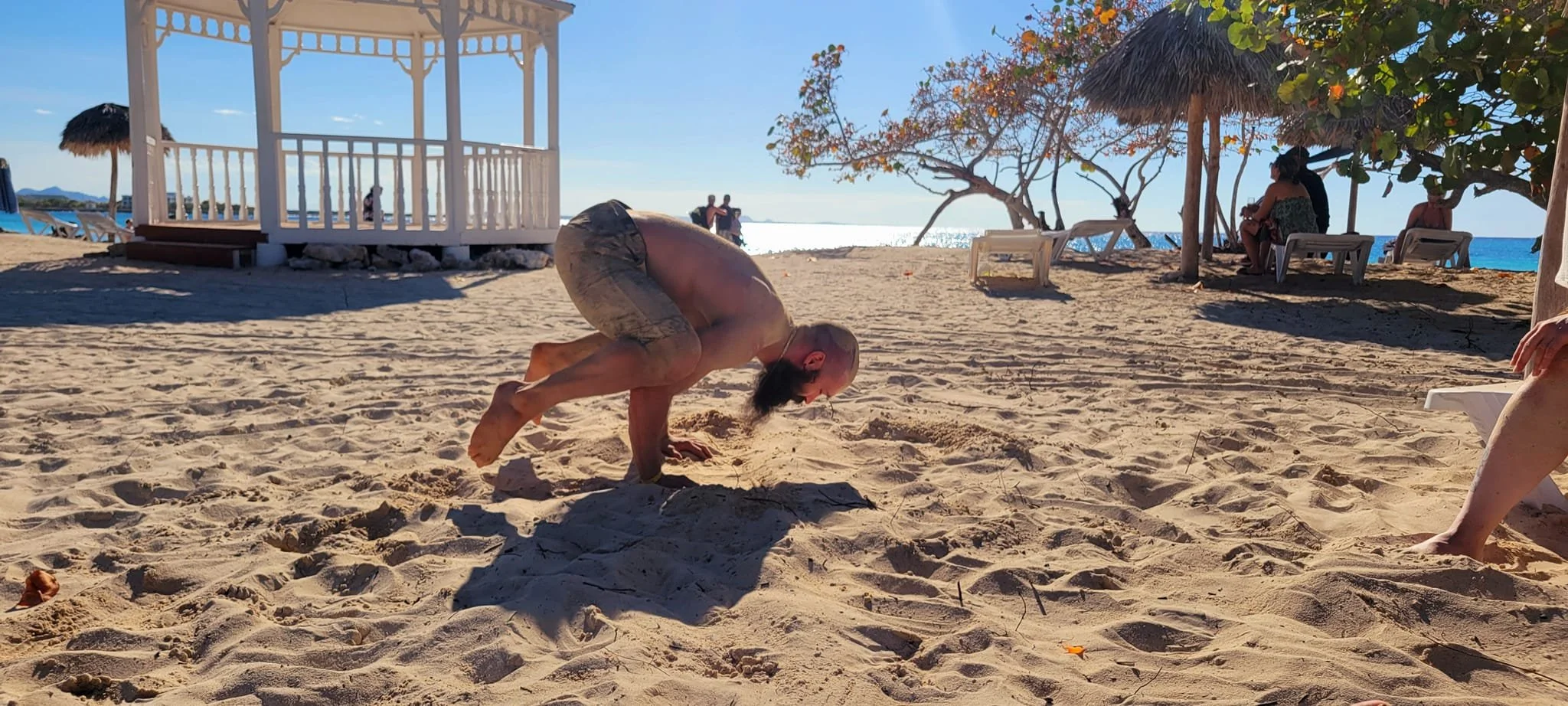 A person doing a handstand on the sandy beach with an ocean view, surrounded by beach umbrellas and chairs, and a gazebo in the background.