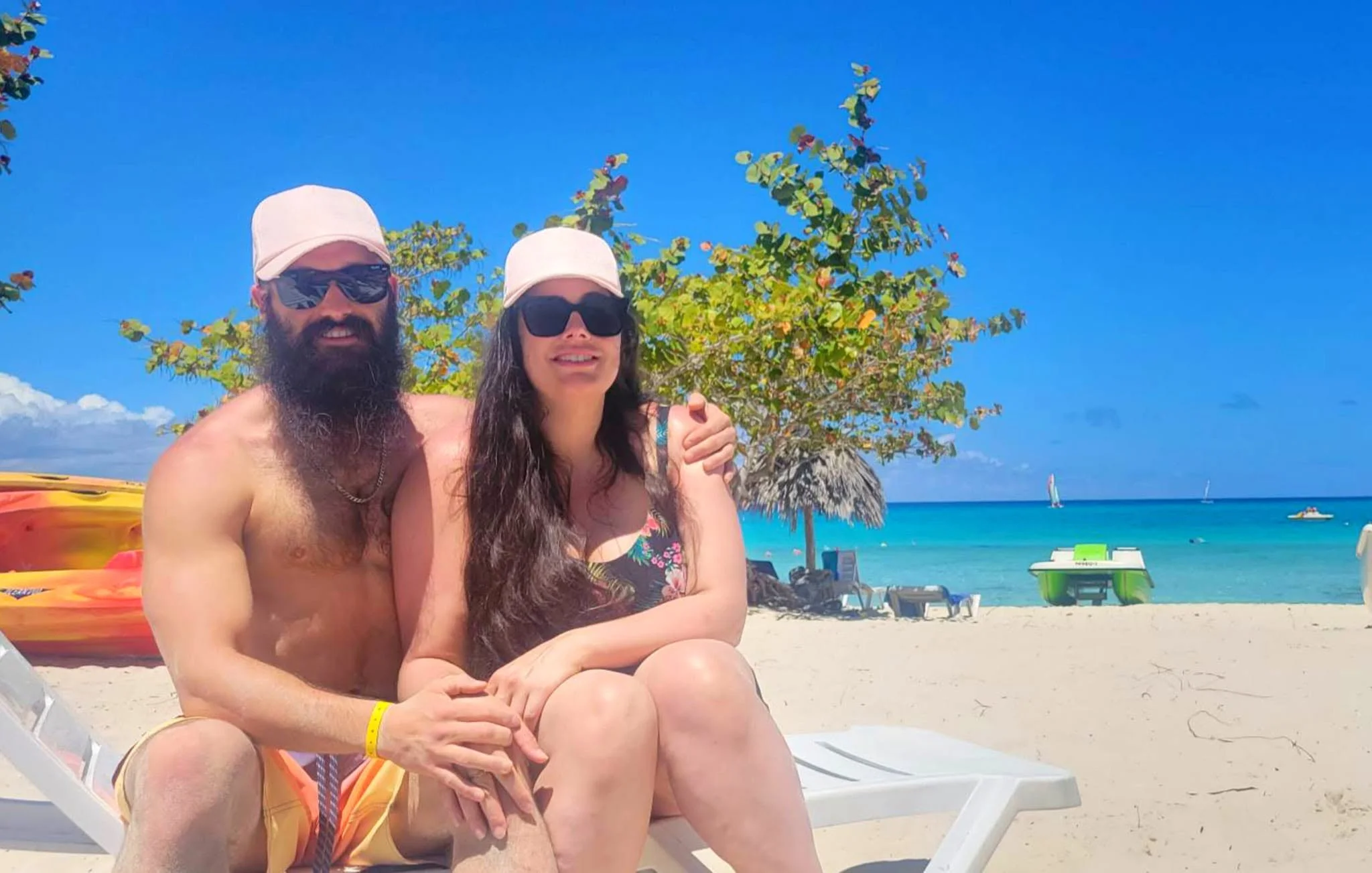 A man and woman sit on a beach chair under a sunny sky. They are wearing sunglasses and hats. The man has a beard and is shirtless, while the woman wears a floral swimsuit. There is a tree behind them and the ocean with sailboats in the background.