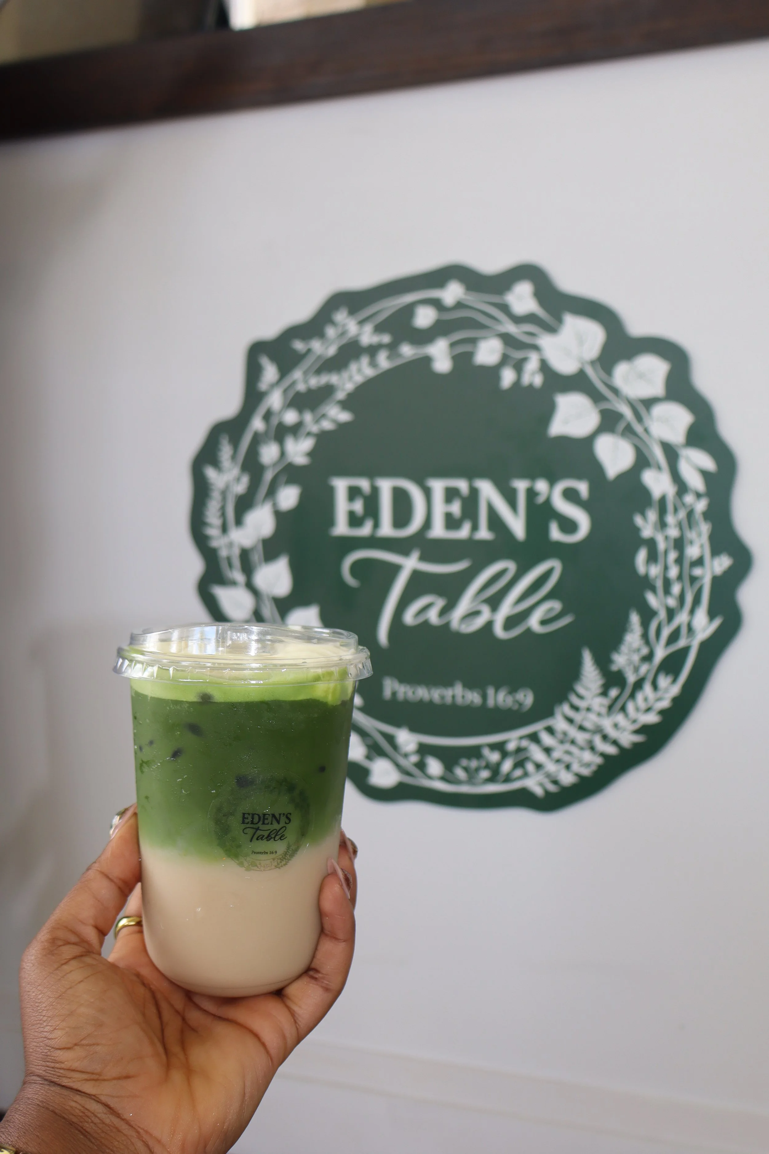 A person holding a cup of matcha green tea latte with cream at Eden's Table, in front of a sign with the location's name and a decorative floral border.