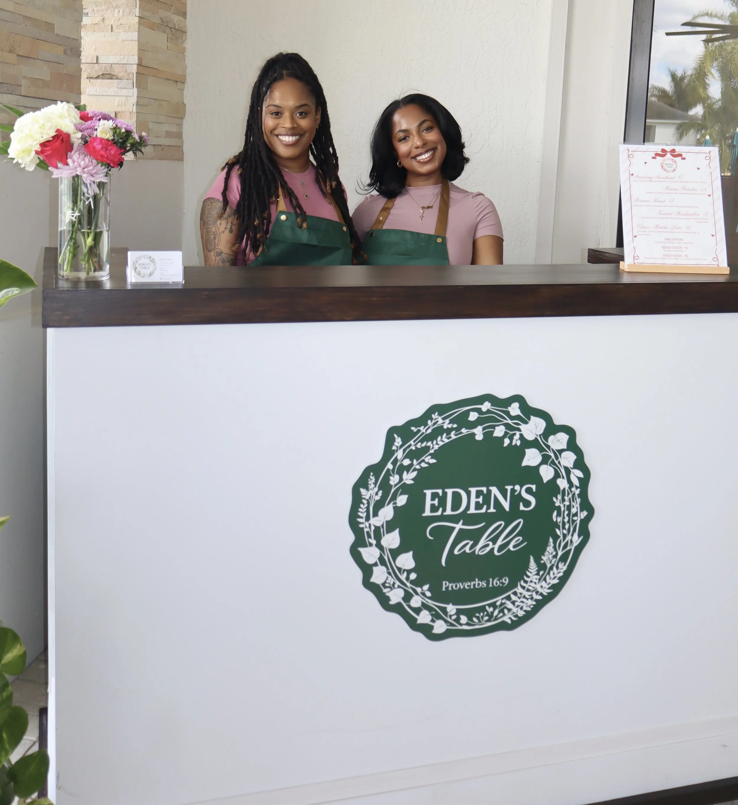 Two women standing behind a reception desk at Eden's Table, smiling, wearing green aprons, with a vase of flowers and a menu on the counter.