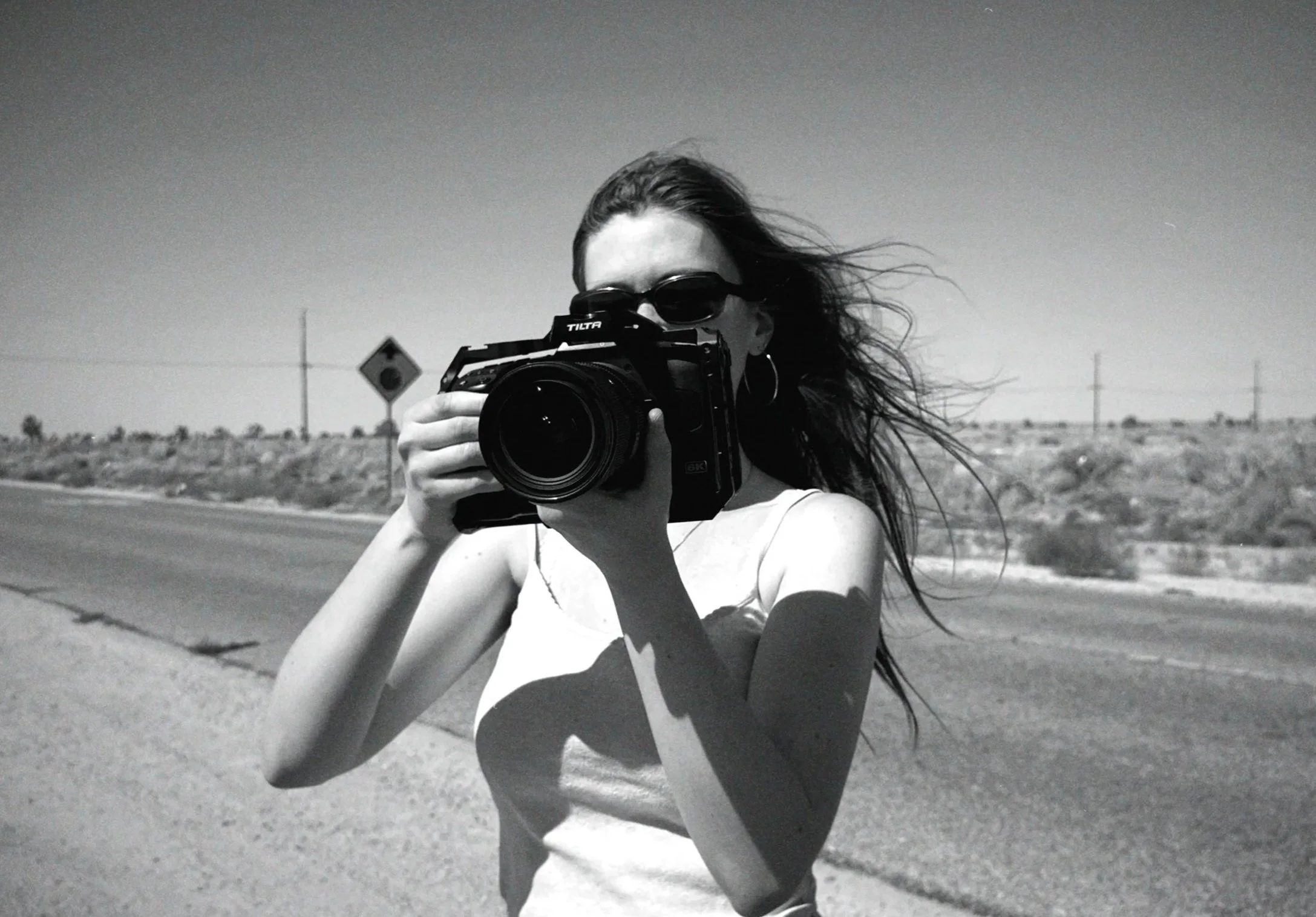 A woman with long hair and sunglasses taking a photo with a camera outdoors on a sunny day, standing on the side of a deserted road with a desert landscape in the background.