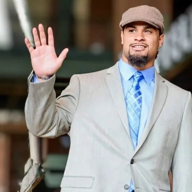 Man in a beige blazer and flat cap waving with a smile