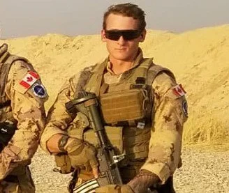 A soldier wearing camouflage uniform, sunglasses, and tactical gear, holding a rifle in a desert-like environment with sand dunes in the background.