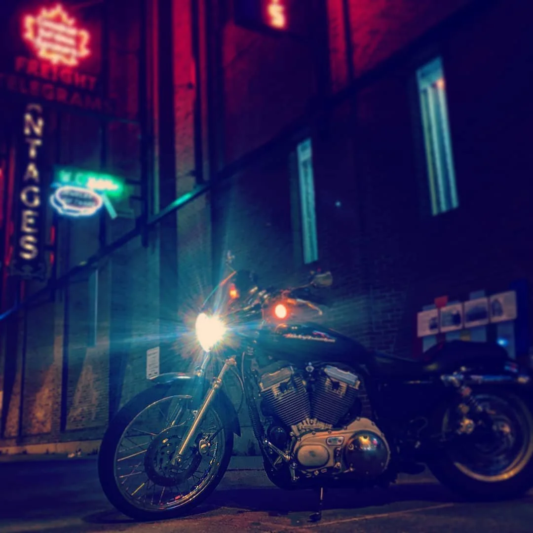 Nighttime scene of a black motorcycle with its headlight on, parked outside a building with illuminated neon signs and brick wall.