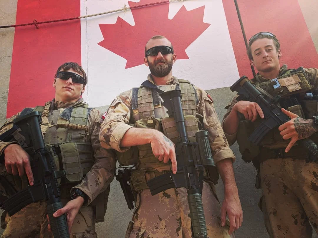 Three soldiers in camouflage military gear standing in front of a Canadian flag.