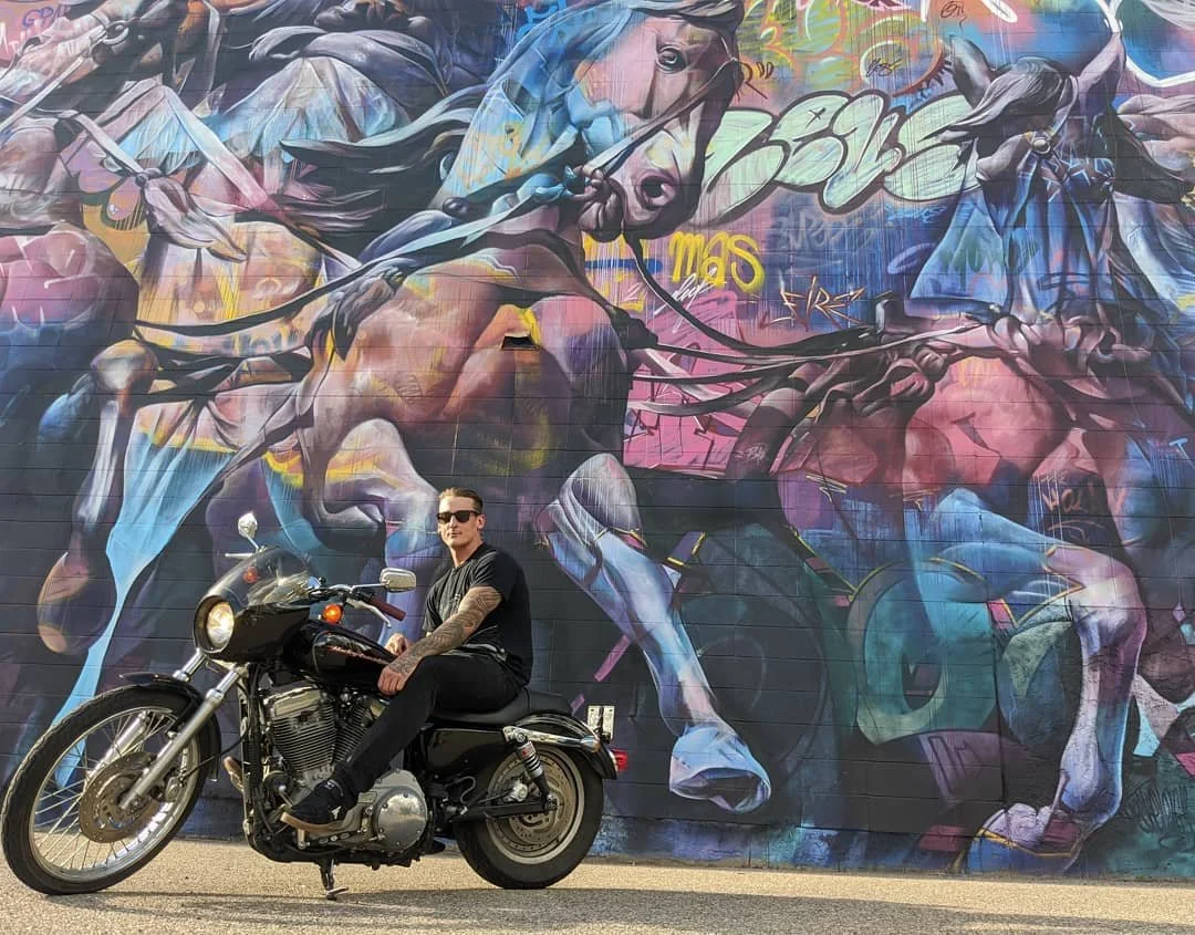 A man with tattoos, wearing sunglasses, a black shirt and black pants, sitting on a black motorcycle in front of a colorful mural of horses painted on a brick wall.
