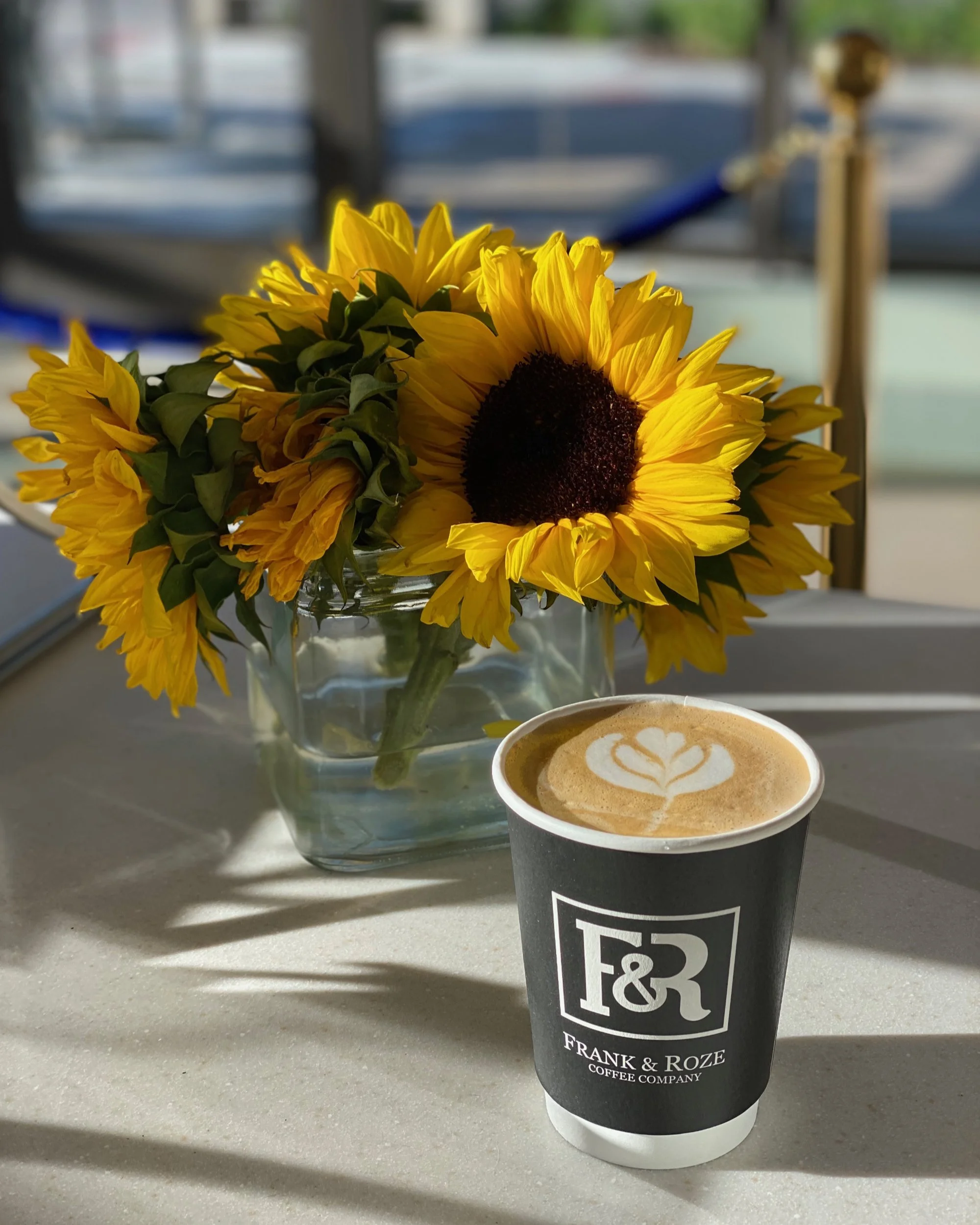 Latté art of a flower in a Frank & Roze to go 12 oz branded coffee cup with a beautiful sunflower arrangement in the background just behind the latté