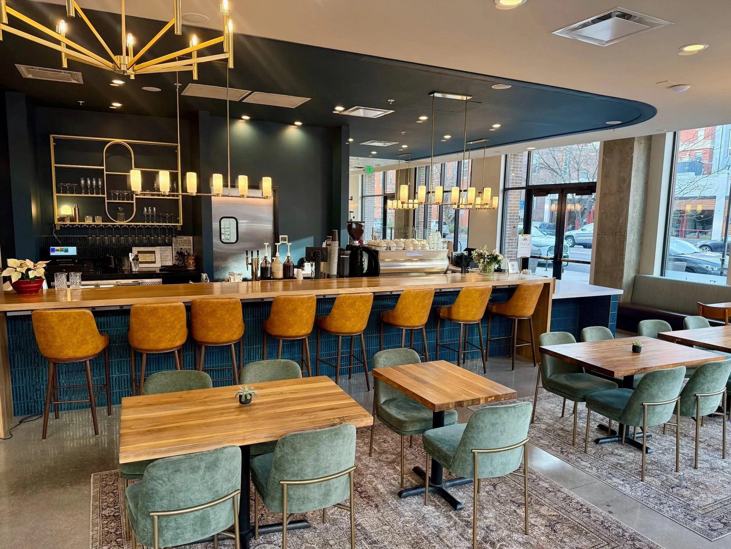 The Uptown Café with modern candle opera matte-brass fixtures, rich teal accents including walls and velvet teal chairs, light brown leather stools against a wood bar with a marble hand off plane in the corner and candle style brass light fixtures ab