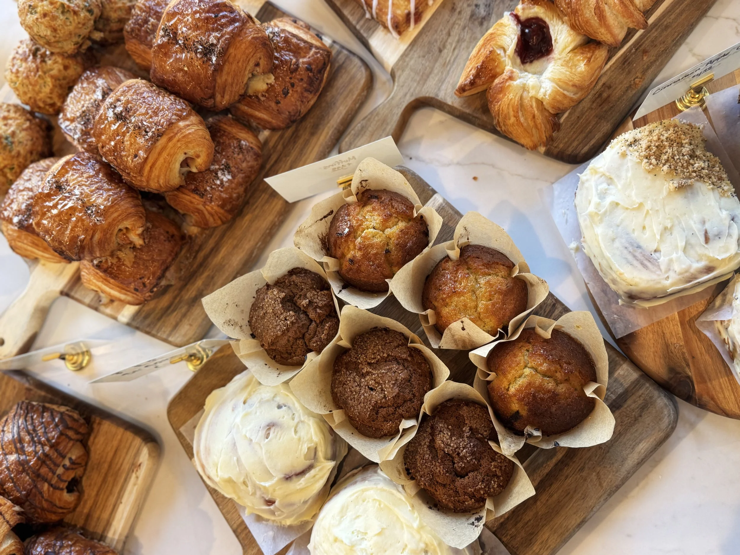 Top down of delicious pastries including muffins, cinnamon rolls, savory ham & guryre pastries, danishes and a lemon swirl roll 