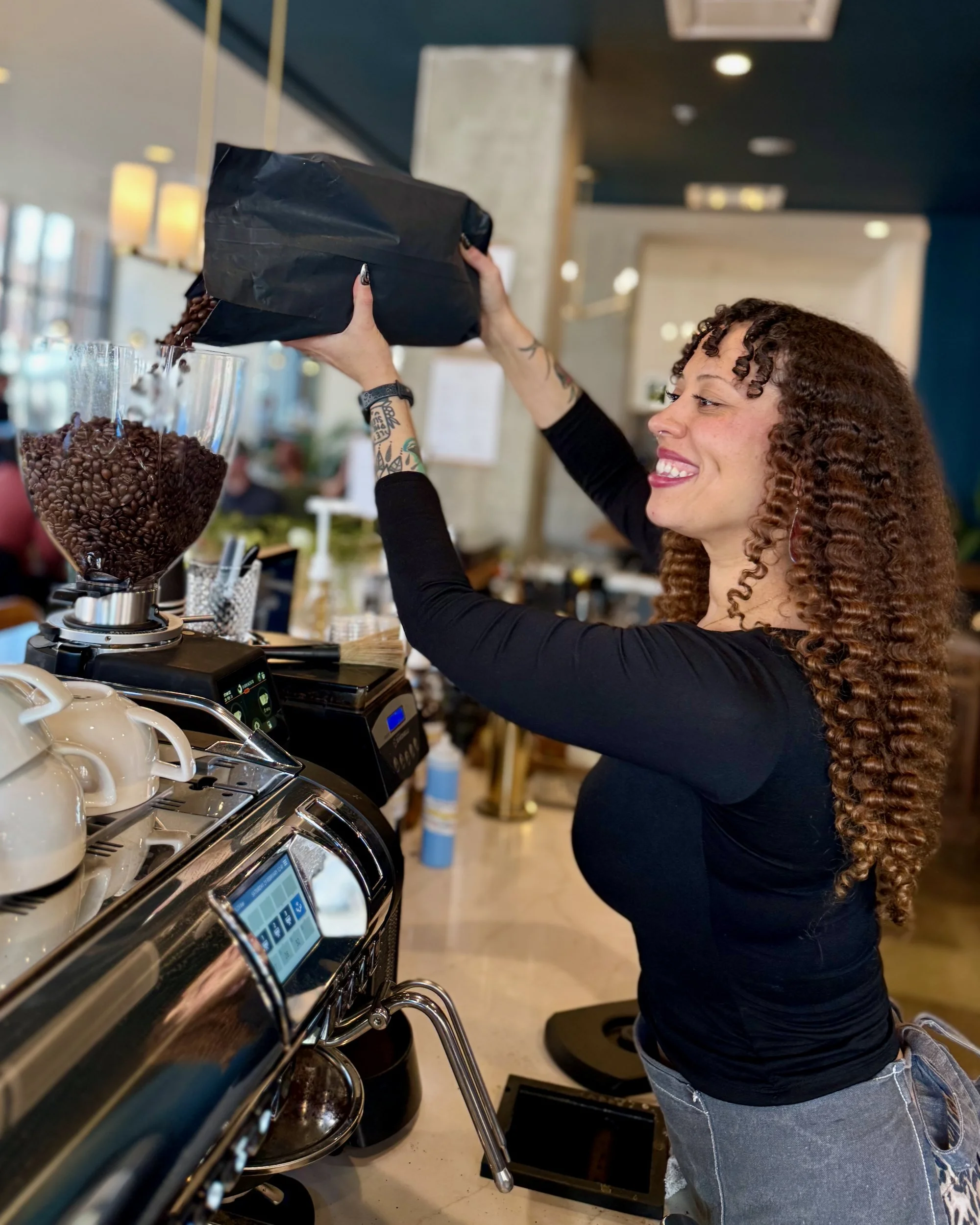 A barista with a big smile and curly long hair pouring coffee beans into the espresso grinder's hopper