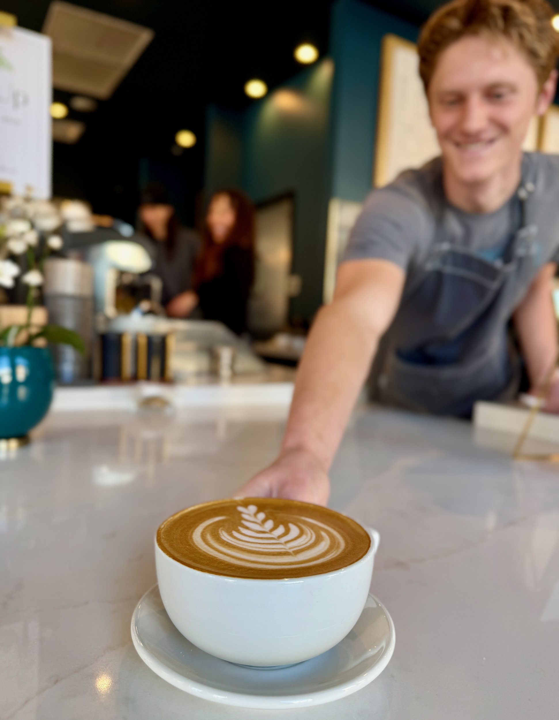A slightly out-of-focus barista smiling as he hands of a latté with beautiful latté art of a small heart with crescents on a white marble hand-off plane.