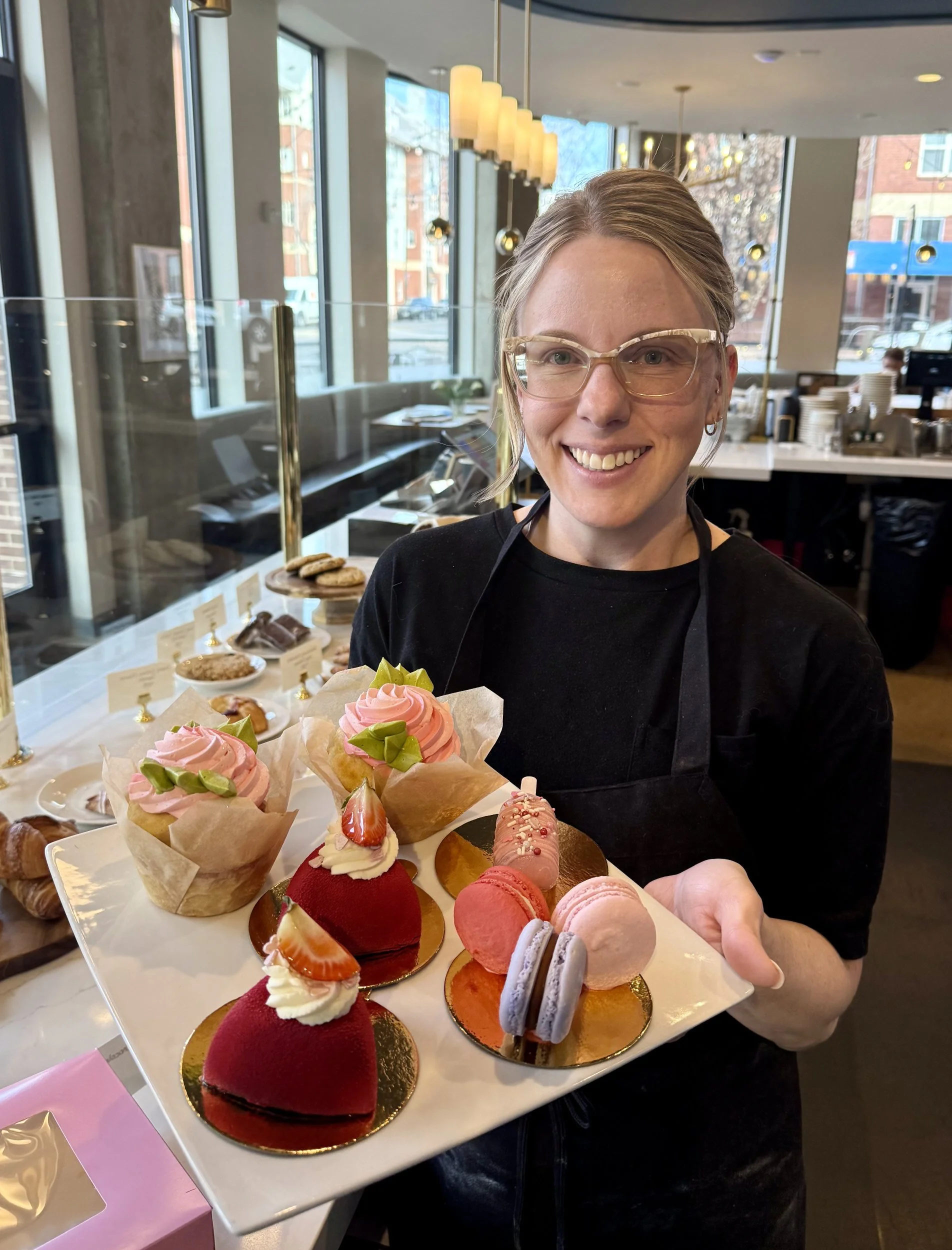 A pastry chef showing off her delicious Valentines Day treats including red velvet  mouse with a strawberry on top, 3 macrons, and 2 pink cup cakes. The café is out of focus in the background