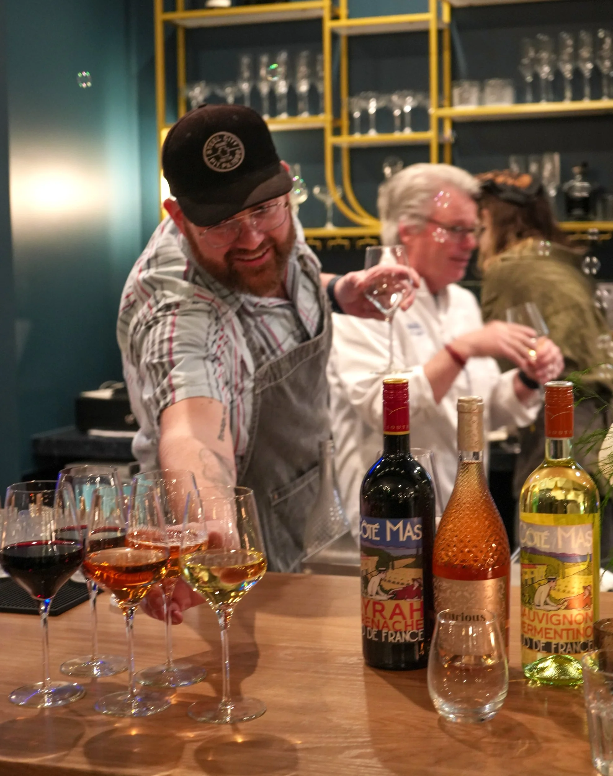 A bar tender with a beard wearing a hat and apron stretching as he pushes a glass of wine across the bar directly at the camera's point of view with Chef Tom in the background. 