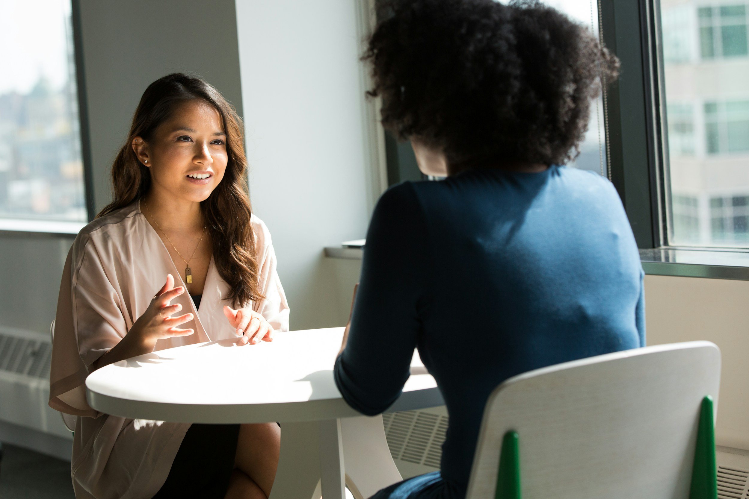 Two women are sitting at a round white table near a window, engaged in a conversation in a well-lit room.