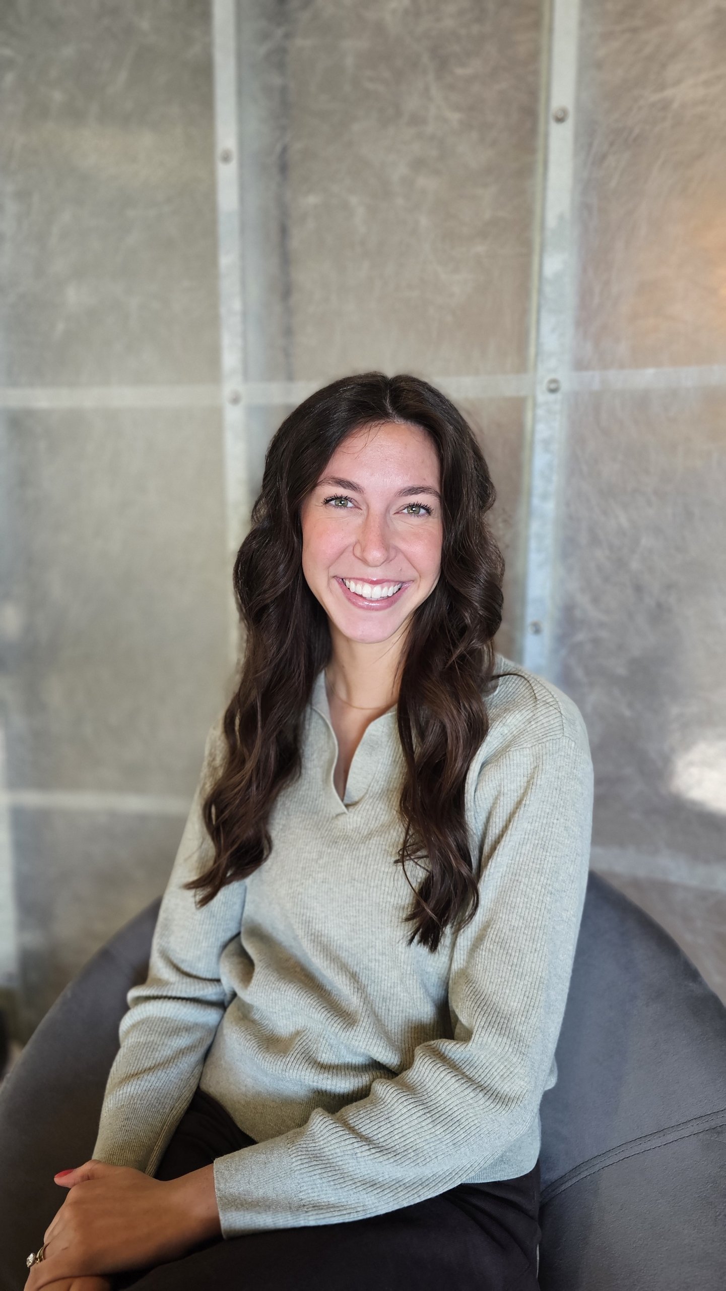 Portrait of a woman with long wavy dark brown hair, light eye color, smiling, wearing a beige sweater, sitting on a gray couch against a textured concrete wall.