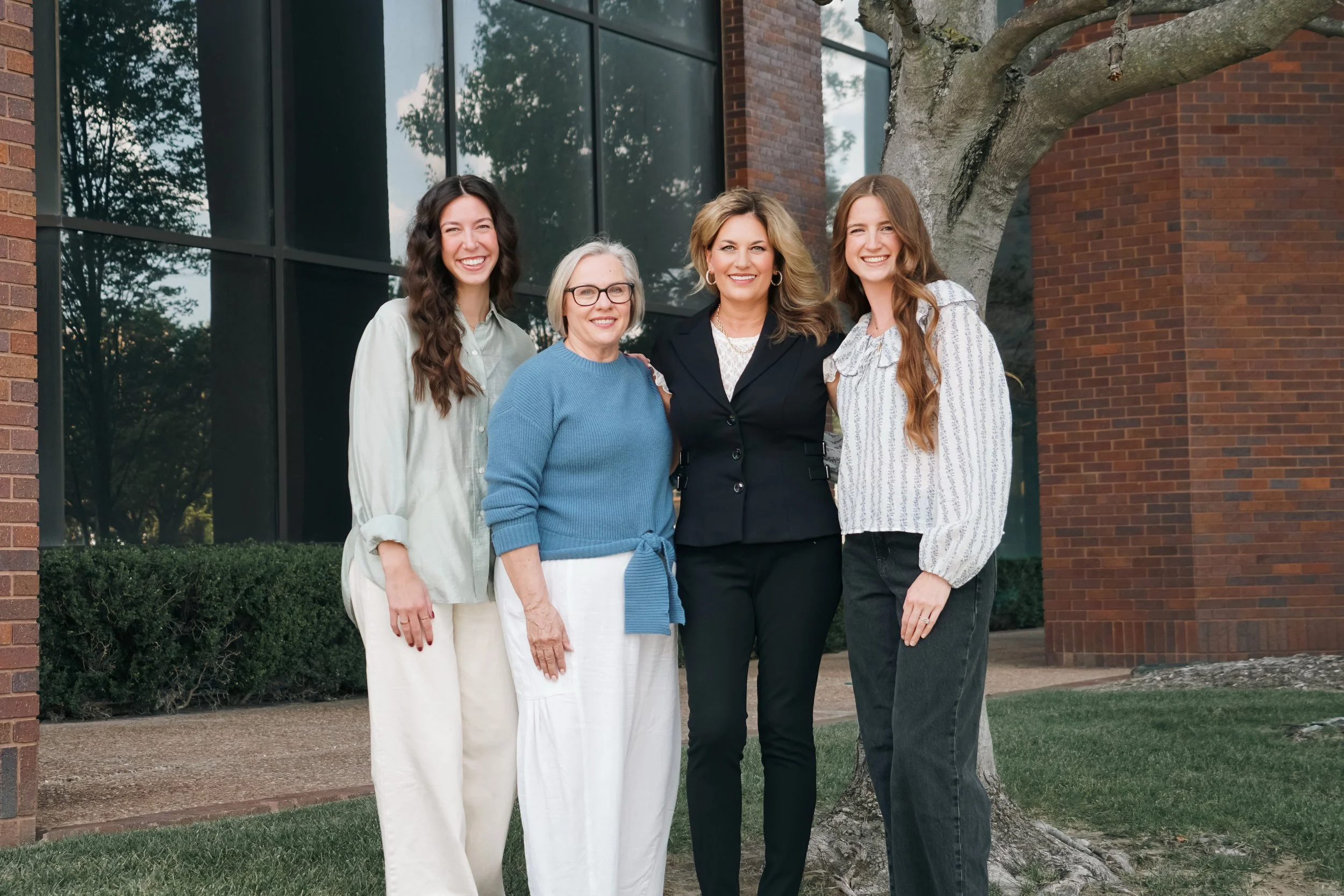 Group of five women standing outside near a brick building and a tree, smiling at the camera.