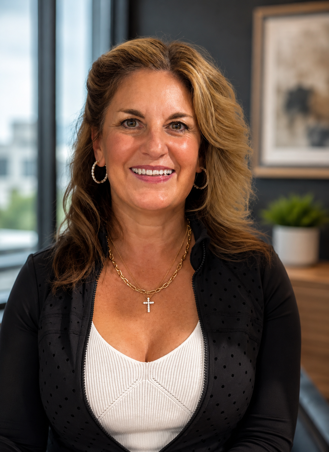 Portrait of a smiling woman with wavy, shoulder-length brown hair, wearing hoop earrings, layered gold necklaces with a cross pendant, a black zip-up jacket with perforations, and a white top, in an indoor setting with large windows and artwork in the background.