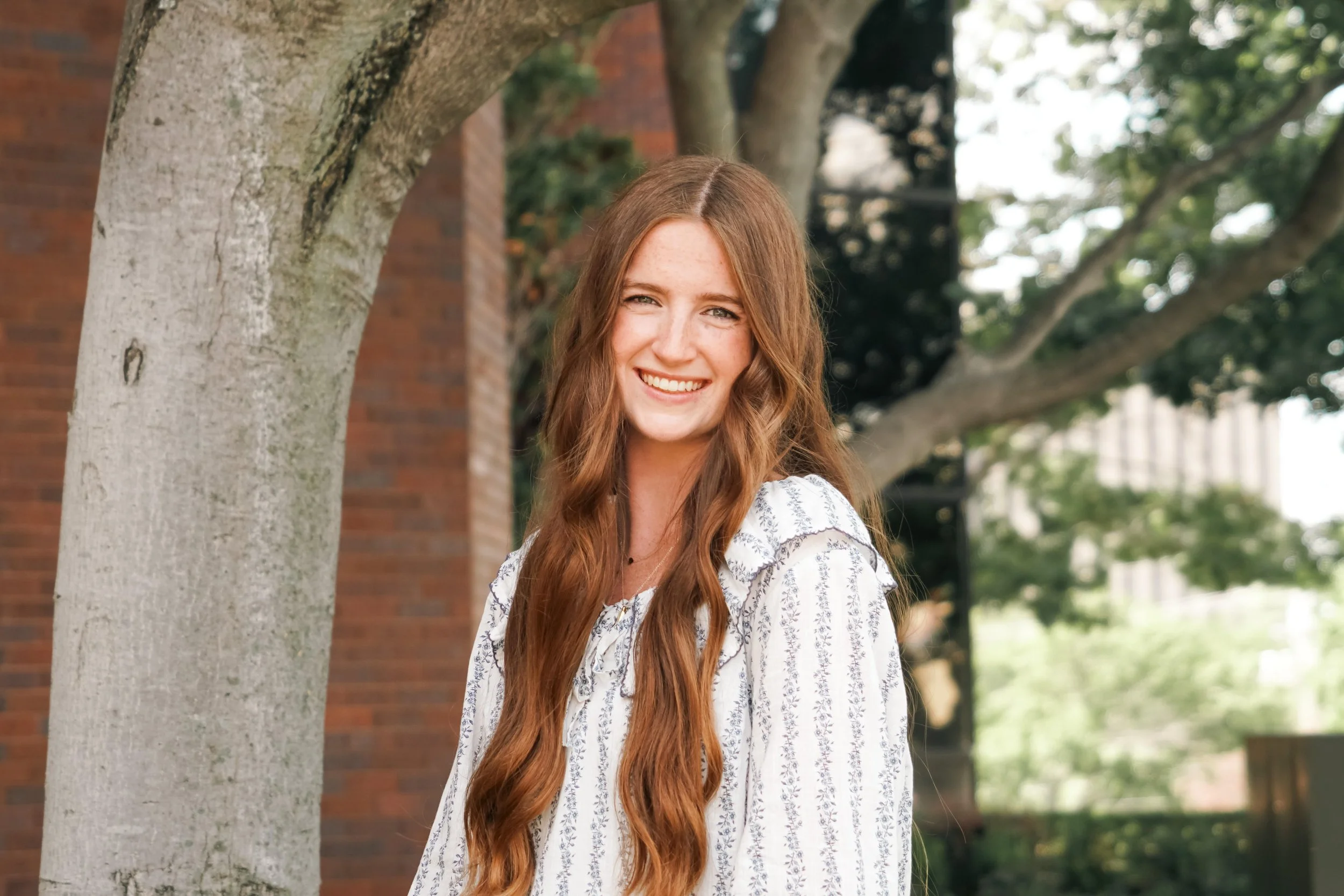 A young woman with long wavy red hair smiling outdoors near a tree. She is wearing a white patterned shirt. The background includes a brick building and green trees.