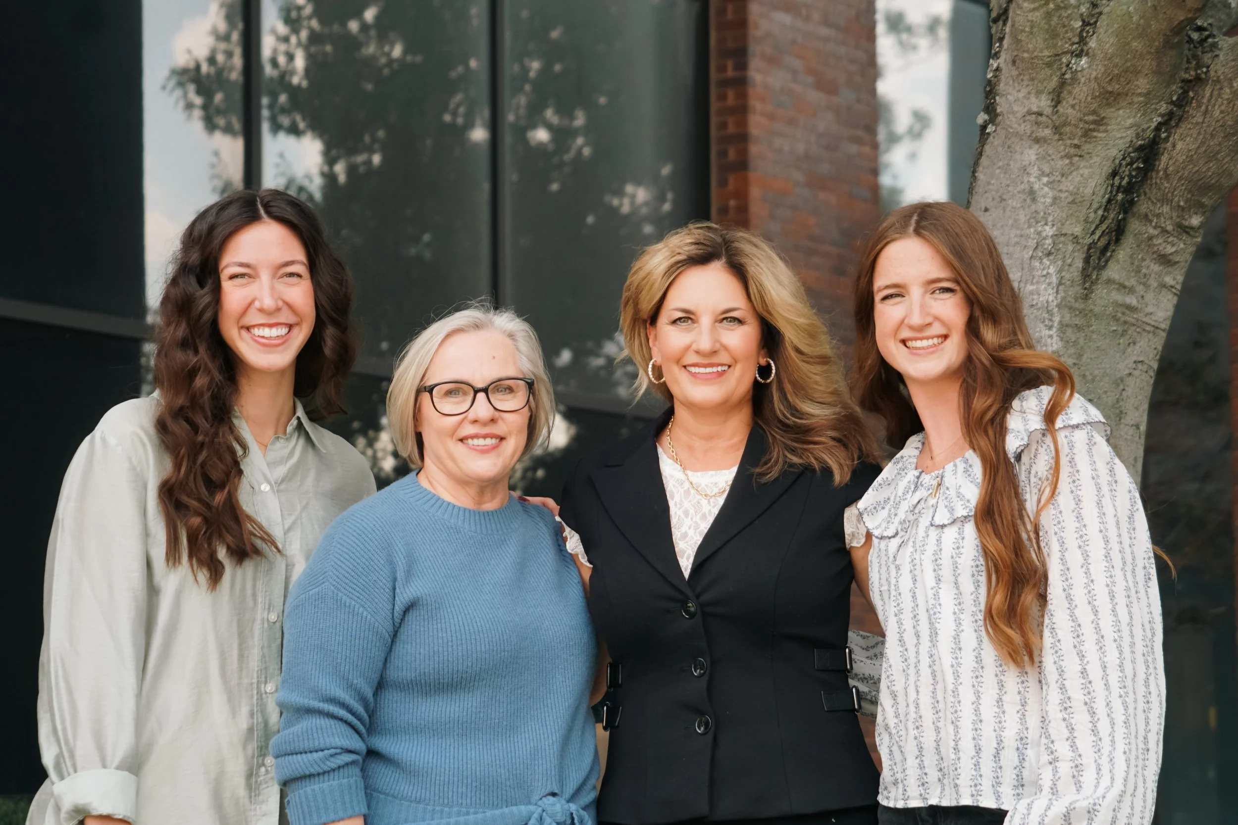 Group of five women smiling outdoors, standing in front of a building with large glass windows, a tree, and brick wall.