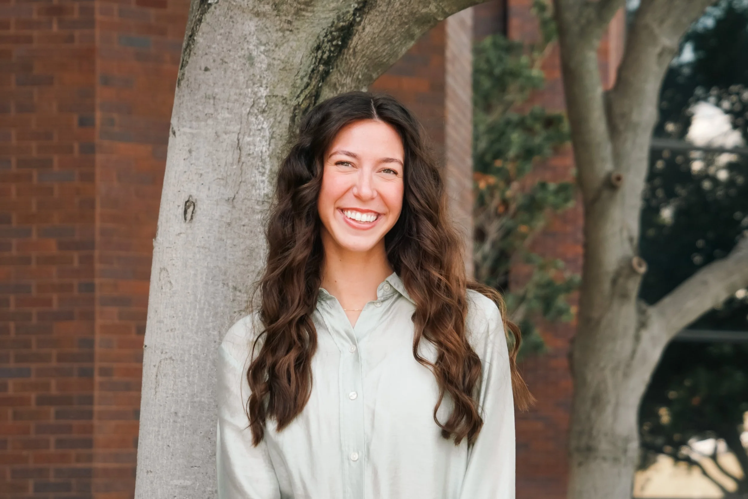 Young woman with long wavy brown hair smiling and standing outdoors next to a tree in front of a brick building.