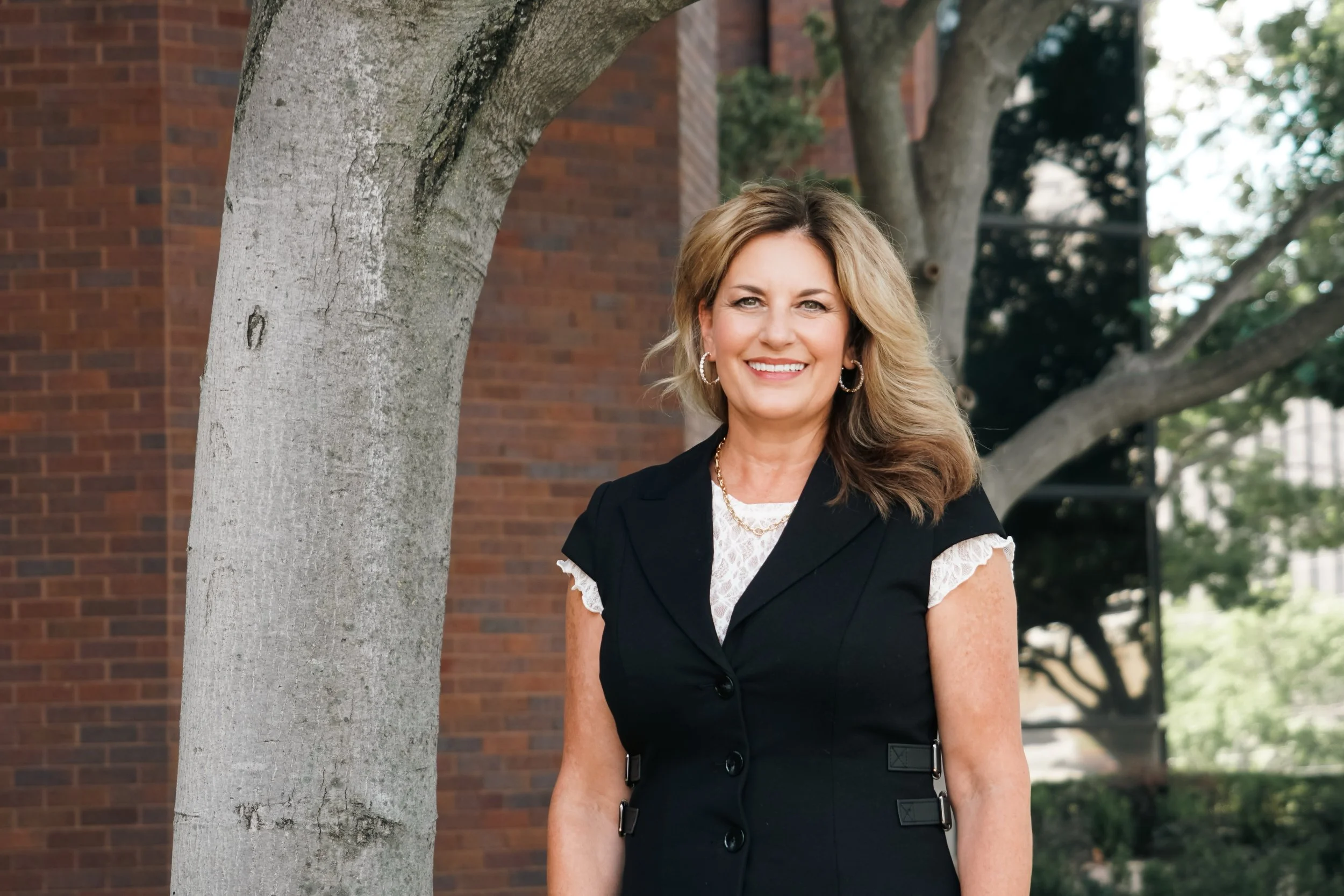 A smiling middle-aged woman with blonde hair, wearing hoop earrings and a black dress with lace sleeves, stands outdoors next to a tree with a brick building in the background.