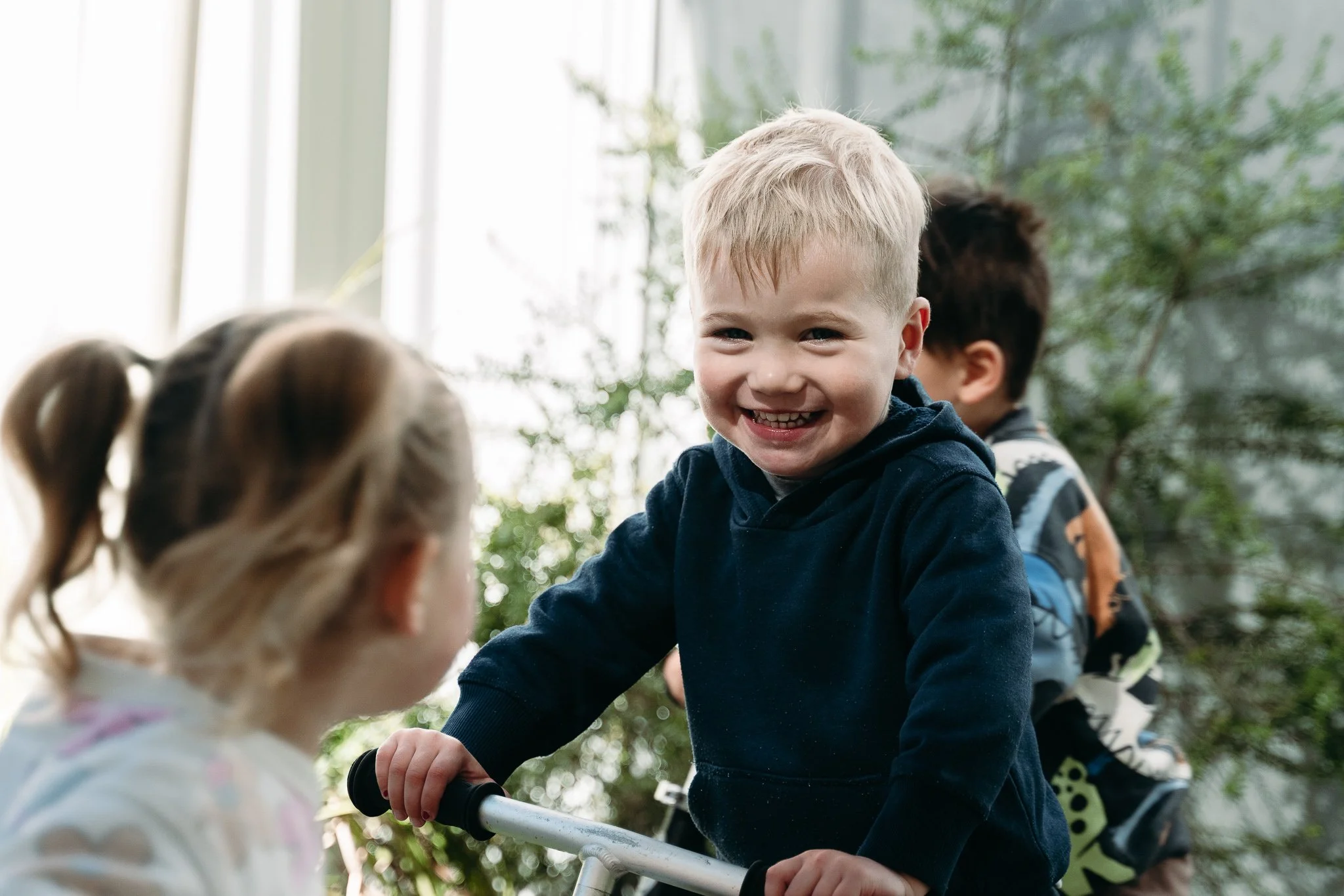 Smiling young boy with blond hair in a dark hoodie playing outdoors with two other children, a girl with pigtails and a boy with dark hair, near green foliage.