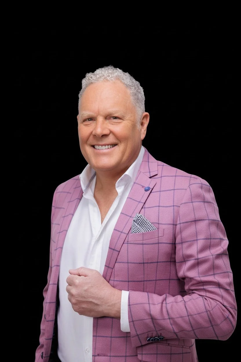 A smiling middle-aged man with curly gray hair wearing a white shirt and a pink checkered blazer with a pocket square, standing against a black background.