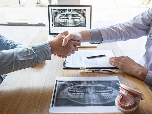 A dentist and a patient shake hands across a table in a dental office. Dental X-rays are visible on a computer monitor and printed on paper, along with a dental model of teeth.