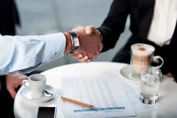 Two people shaking hands over a table with documents, a coffee cup, a glass of water, an iced coffee, a smartphone, and a pen.